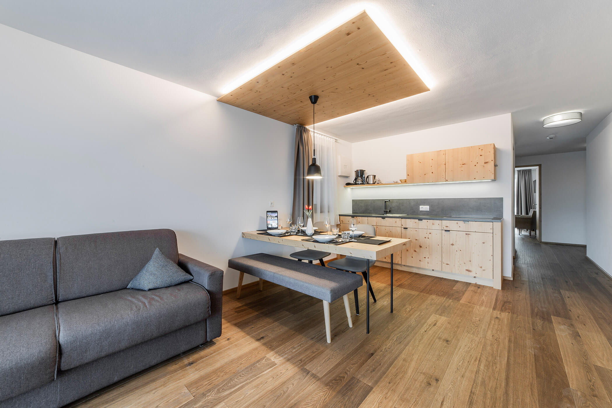 Appartamento moderno con pavimenti in legno, un divano grigio, un tavolo da pranzo e una cucina con mobili in legno. - Hotel Oberwiesen