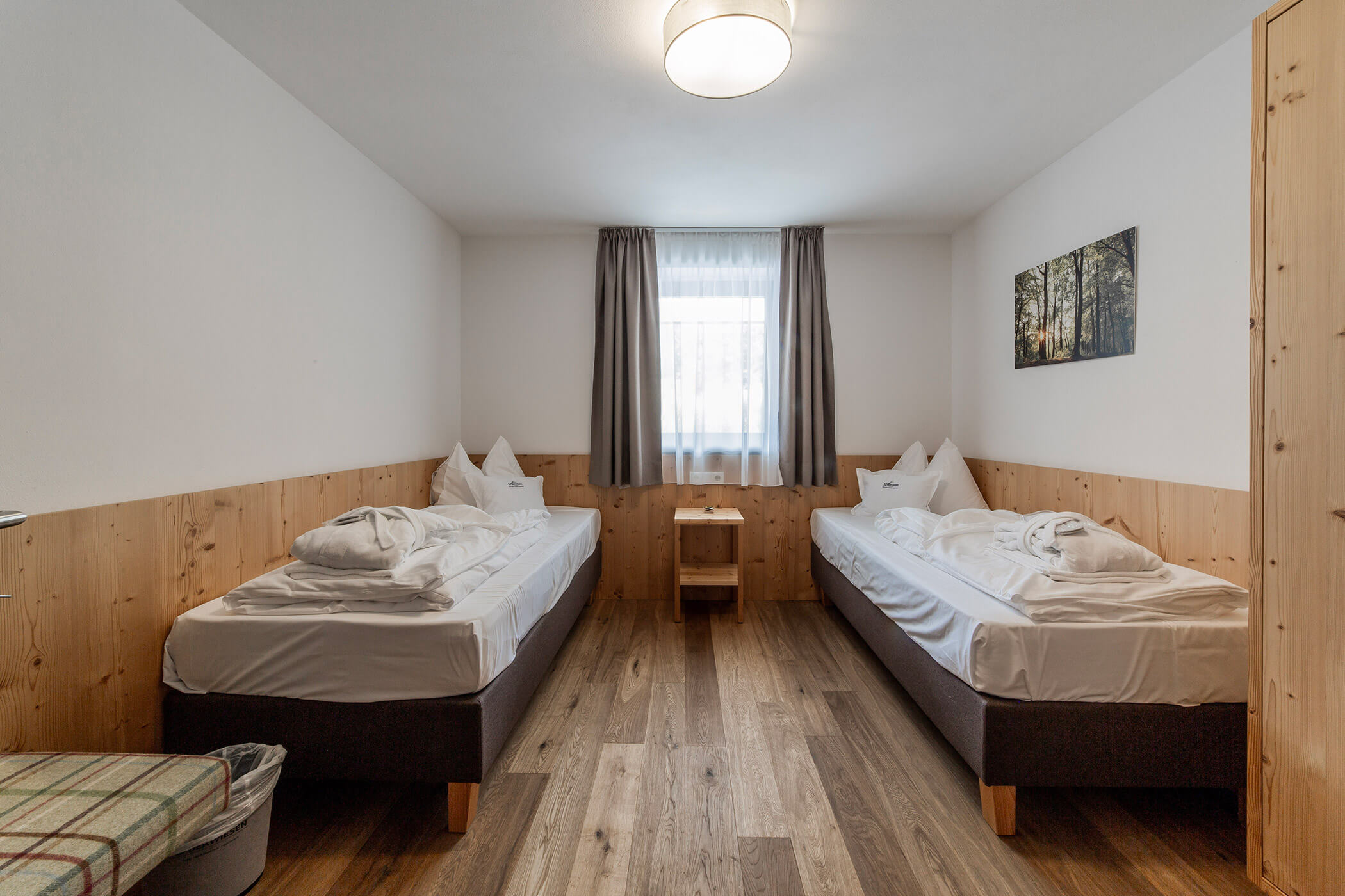 Due letti singoli in una stanza semplice e ordinata, con rivestimenti in legno, una finestra e un quadro raffigurante un bosco appeso alla parete. - Hotel Oberwiesen