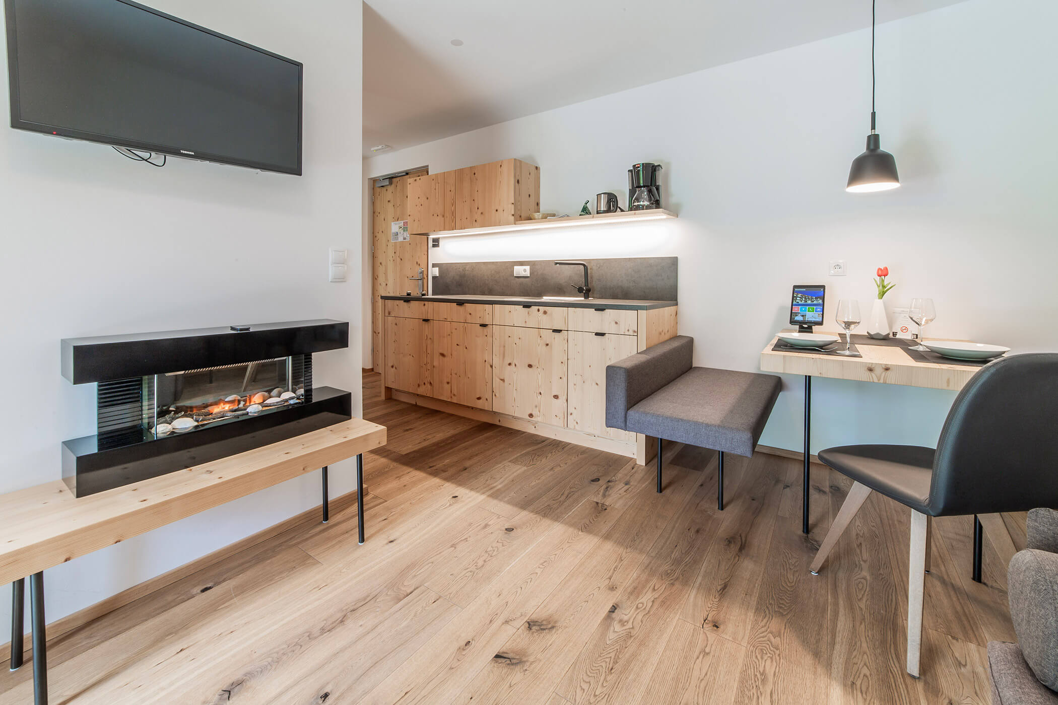 Modernes Studio mit Holzakzenten, Kochnische, an der Wand montiertem Fernseher, elektrischem Kamin und Essecke. - Hotel Oberwiesen