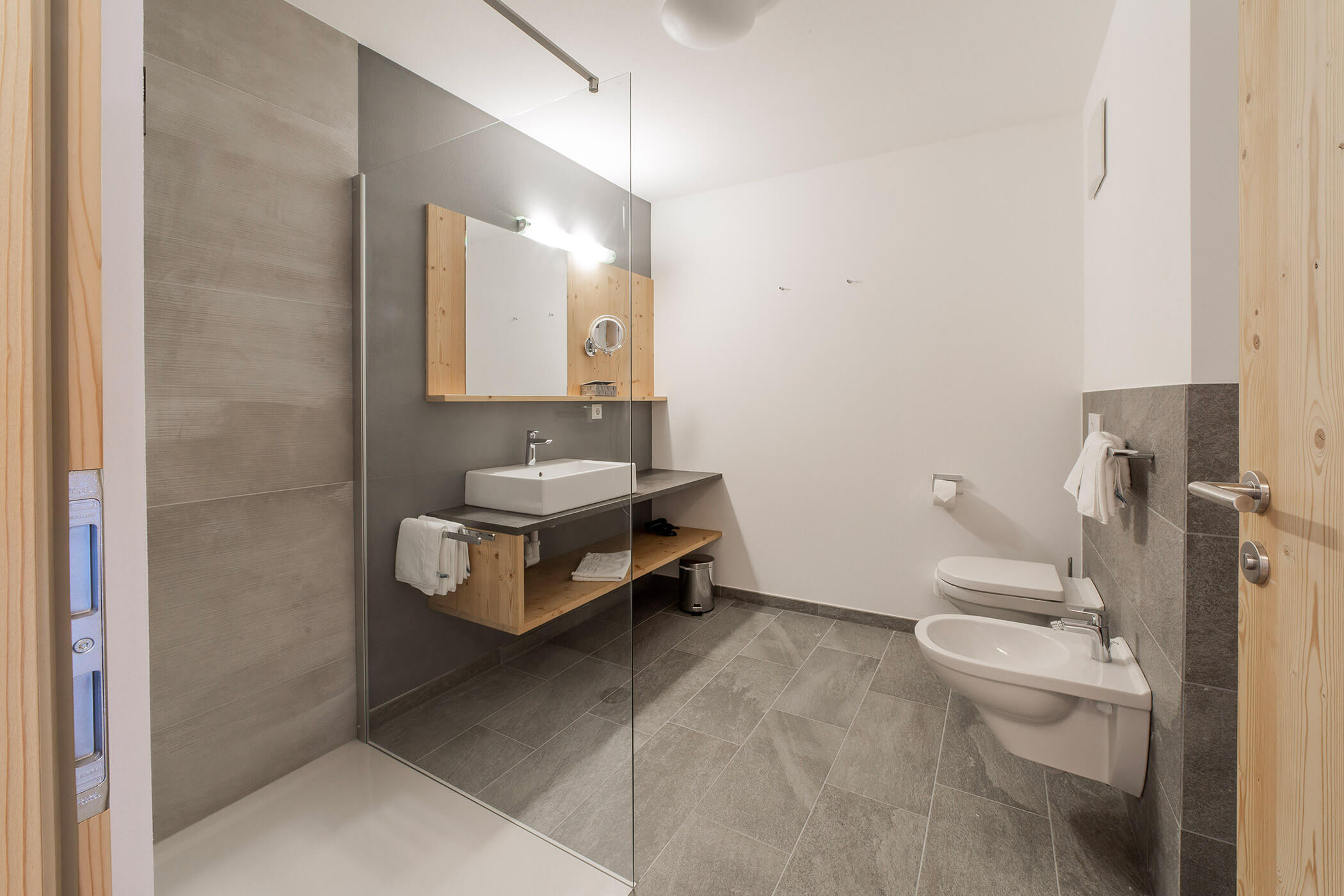 Bagno moderno con cabina doccia, lavabo, specchio, bidet e WC, il tutto su un pavimento piastrellato grigio. - Hotel Oberwiesen