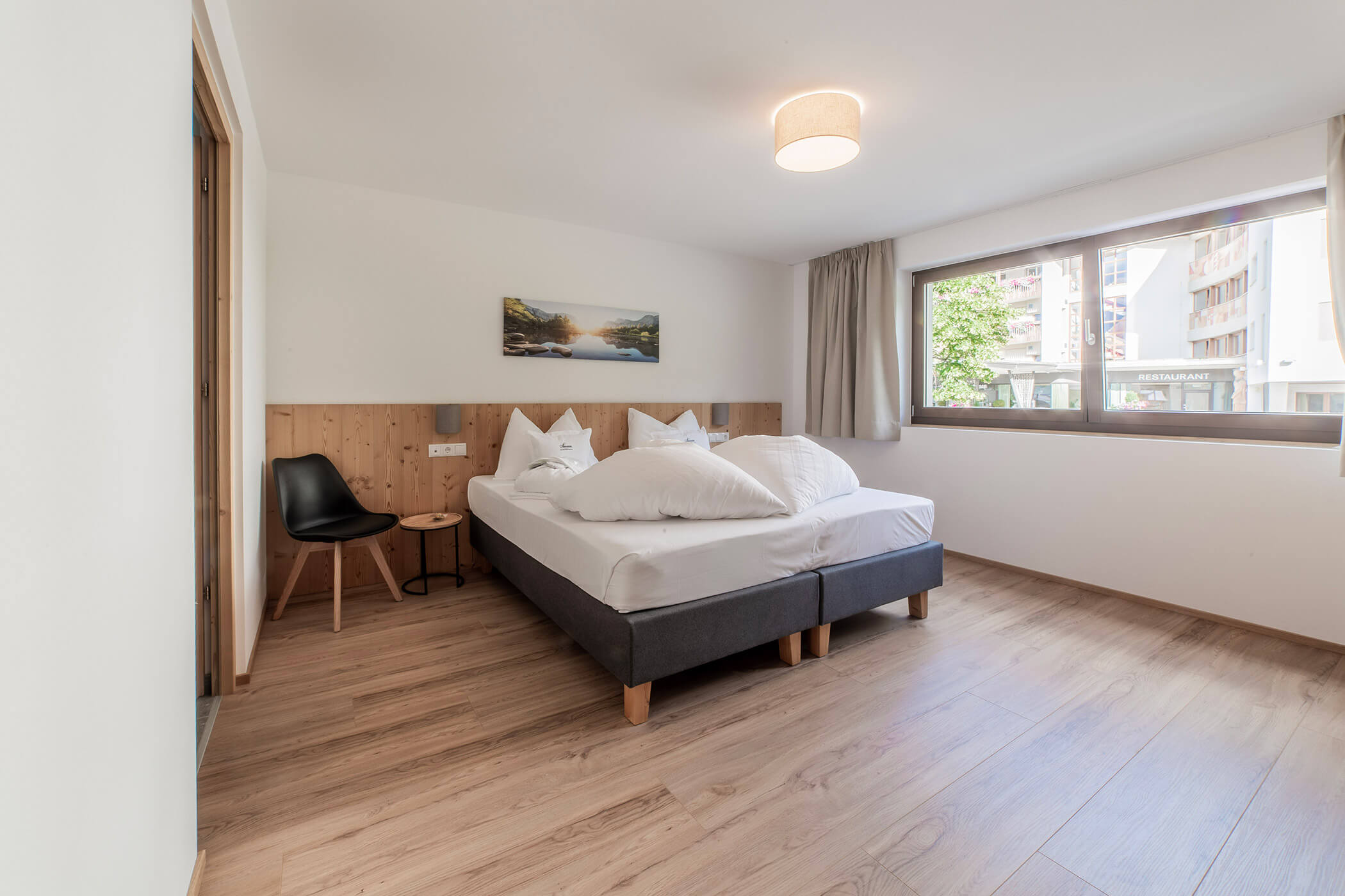 Camera da letto luminosa con un letto matrimoniale, pavimenti in legno, una sedia, opere d’arte e una grande finestra. - Hotel Oberwiesen