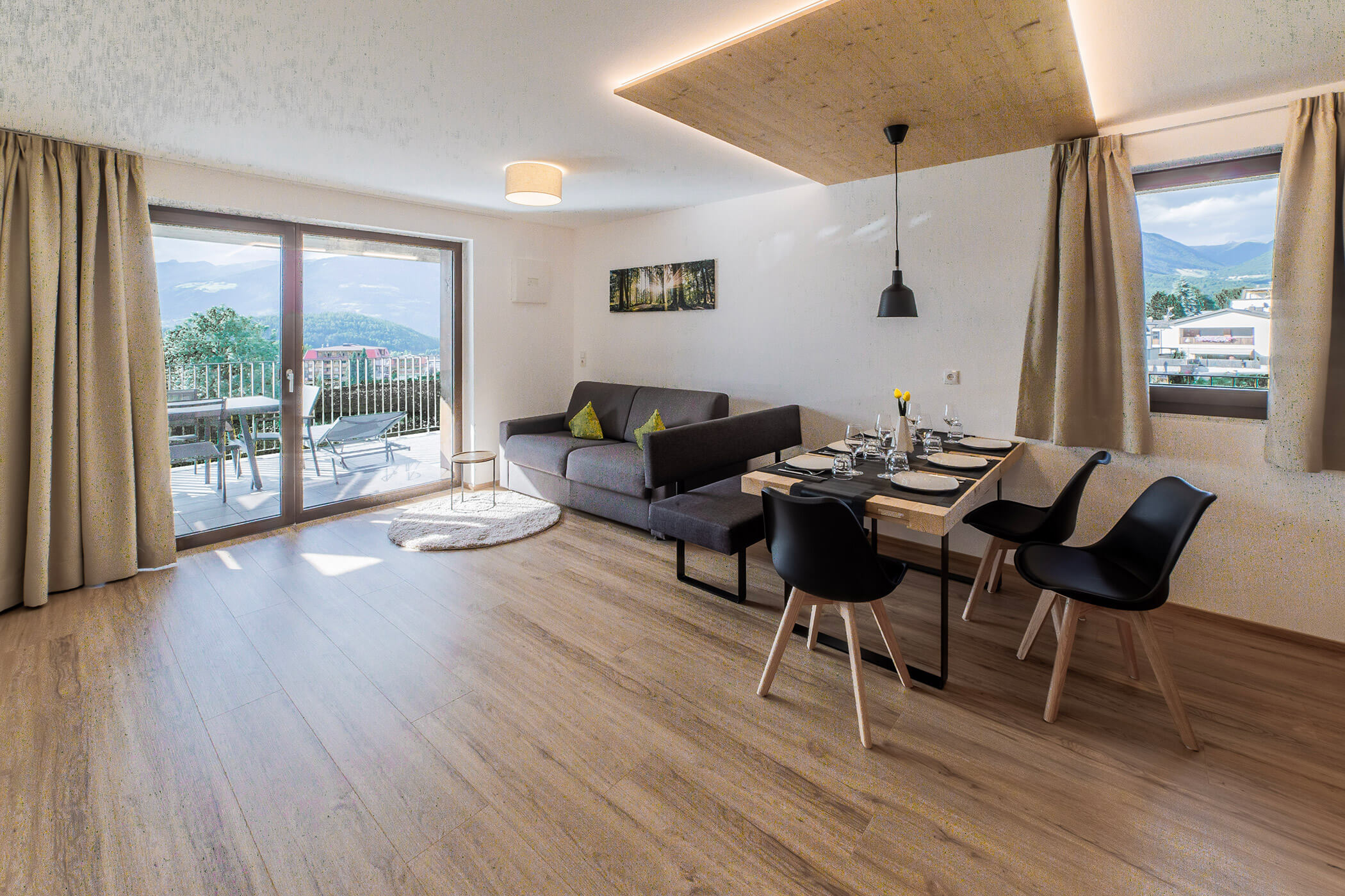 Moderno soggiorno-sala da pranzo con pavimenti in legno, divano grigio, tavola imbandita, ampie finestre e vista sulle montagne. - Hotel Oberwiesen