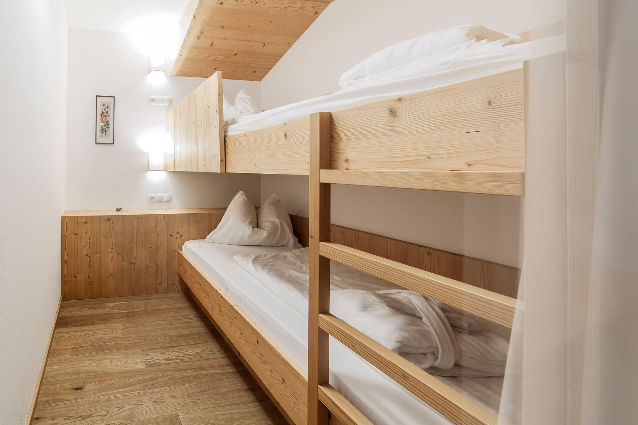 Piccola stanza con letti a castello in legno, biancheria da letto bianca, pavimento in legno, applique e un quadro incorniciato. - Hotel Oberwiesen