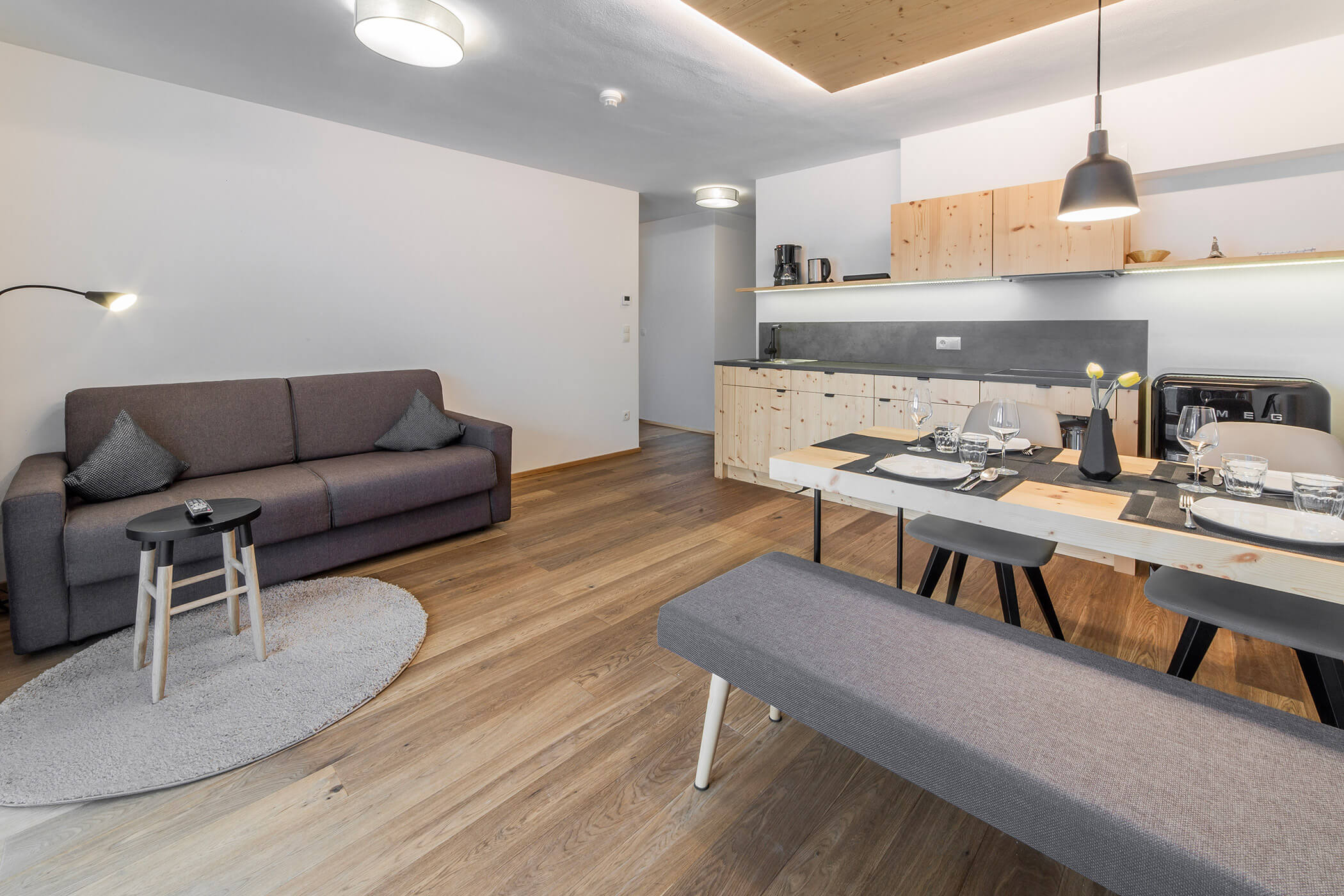 Moderna zona soggiorno-pranzo con divano grigio, tavolo in legno, panca e arredamento minimalista. - Hotel Oberwiesen