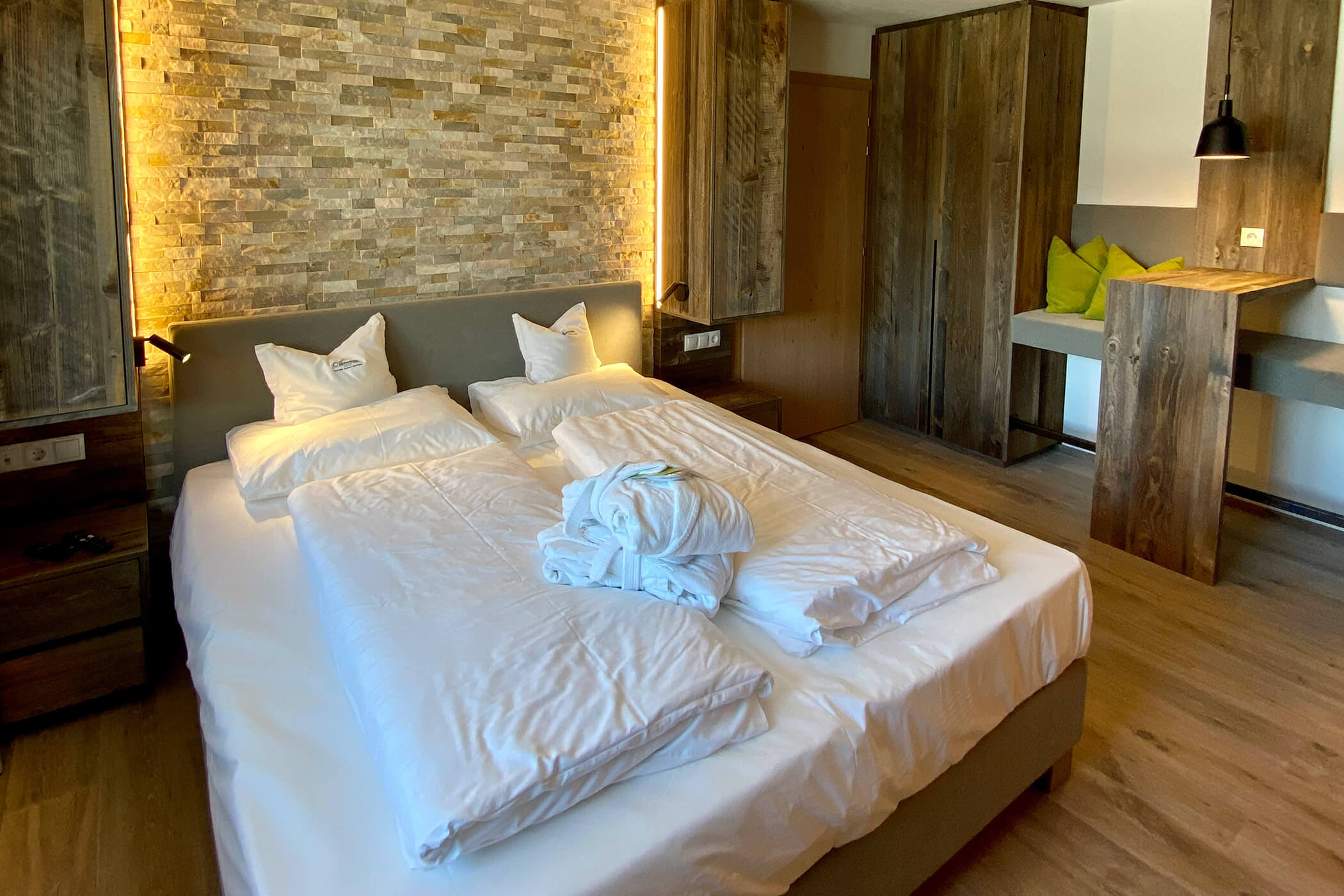 Camera d'albergo moderna con letto matrimoniale, parete con inserti in pietra, mobili in legno e accappatoi ripiegati sul letto. - Hotel Oberwiesen
