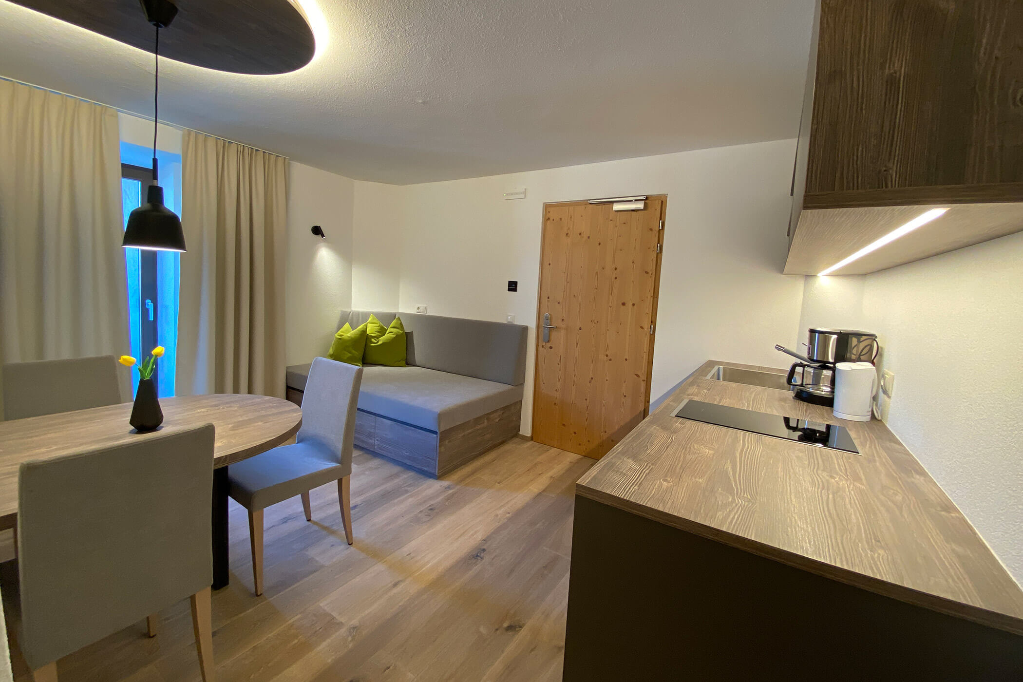 Cucina moderna e zona pranzo con mobili in legno, divano grigio e cuscini verdi vicino a una porta in legno. - Hotel Oberwiesen