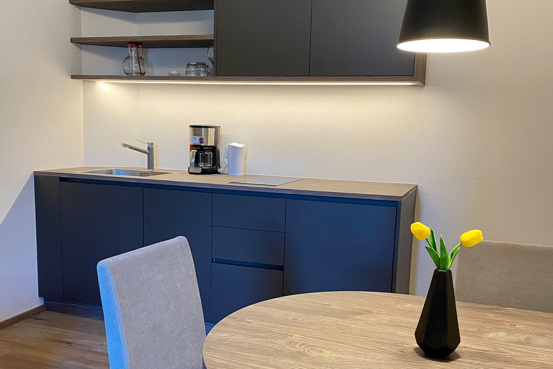 Minimalistische Küche mit dunklen Schränken, einer Kaffeemaschine und einem Tisch mit einer Vase, in der gelbe Tulpen stehen. - Hotel Oberwiesen