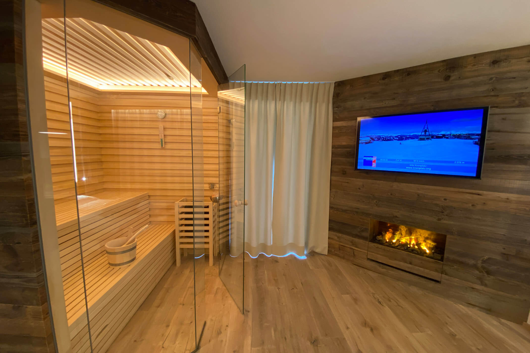 Camera moderna con sauna in vetro, pareti in legno, caminetto, TV e tende bianche alla finestra. - Hotel Oberwiesen
