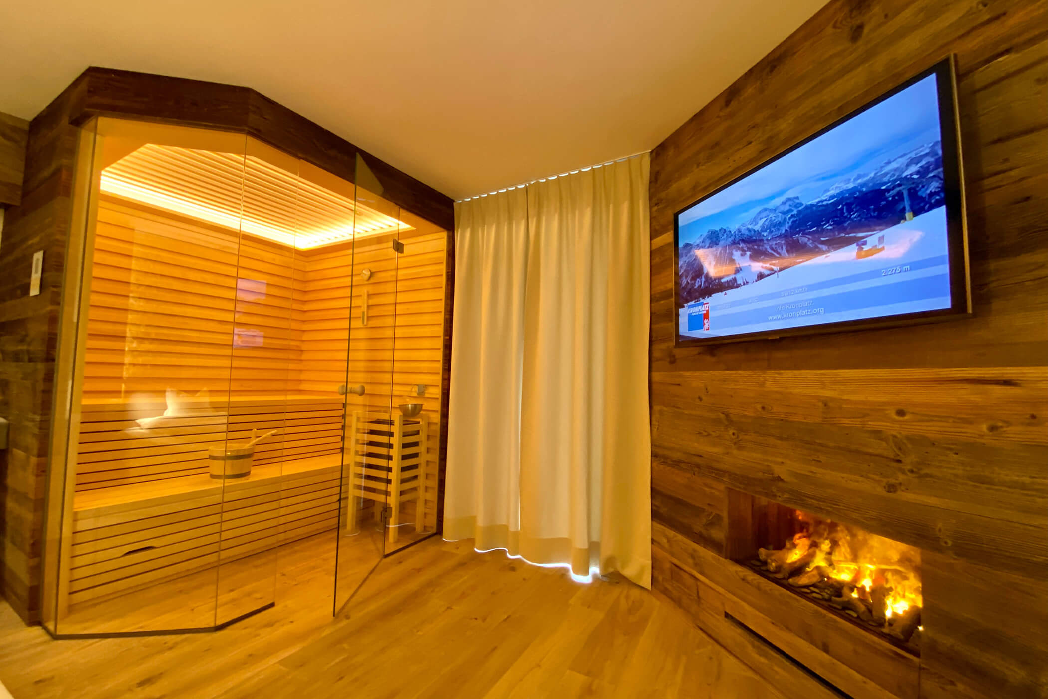 Camera moderna con sauna in vetro, pareti in legno, caminetto, TV con immagini di montagne e tenda che copre la finestra. - Hotel Oberwiesen