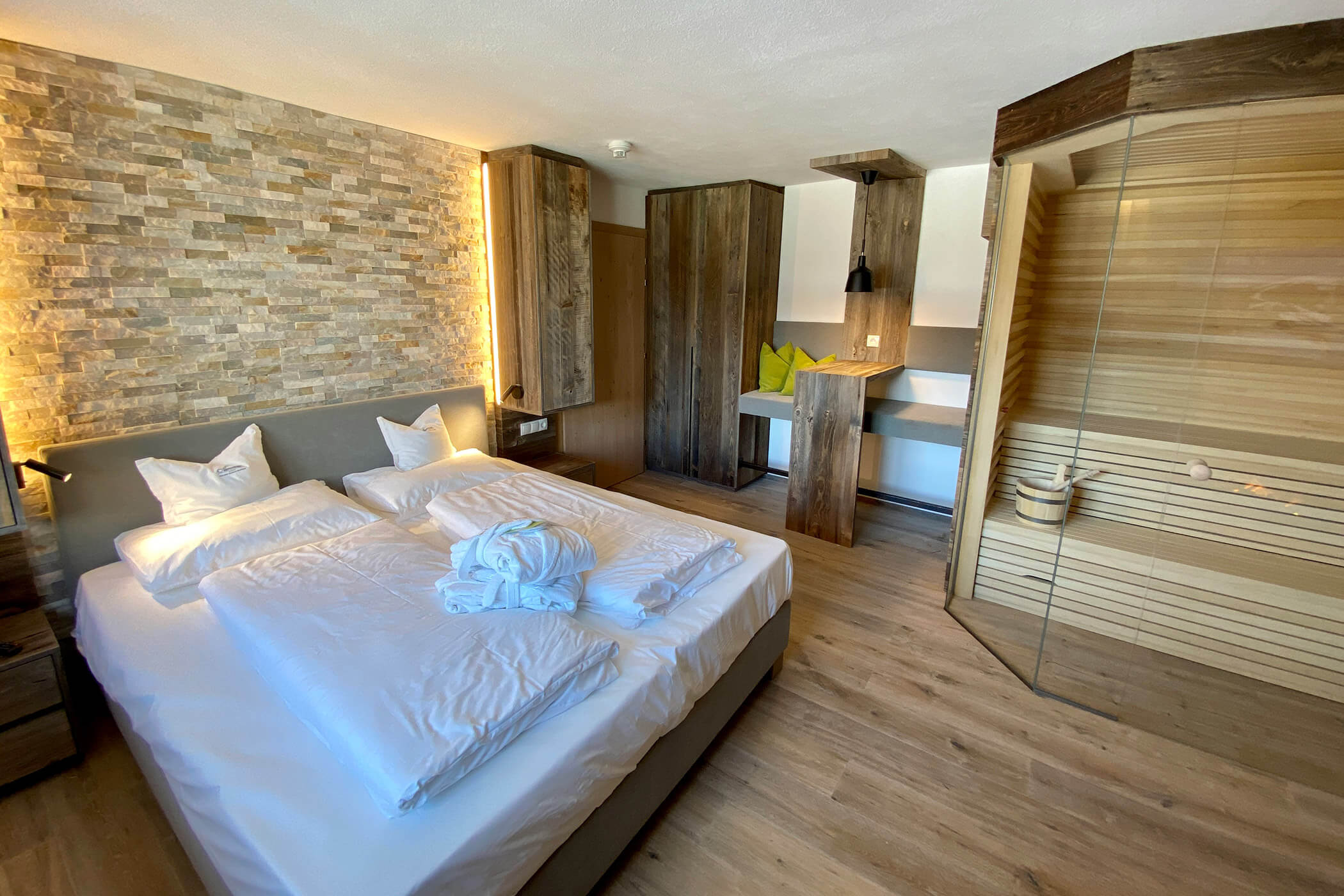 Moderna camera d'albergo con letto matrimoniale, parete decorativa in pietra, angolo cottura e sauna con pareti in vetro sulla destra. - Hotel Oberwiesen