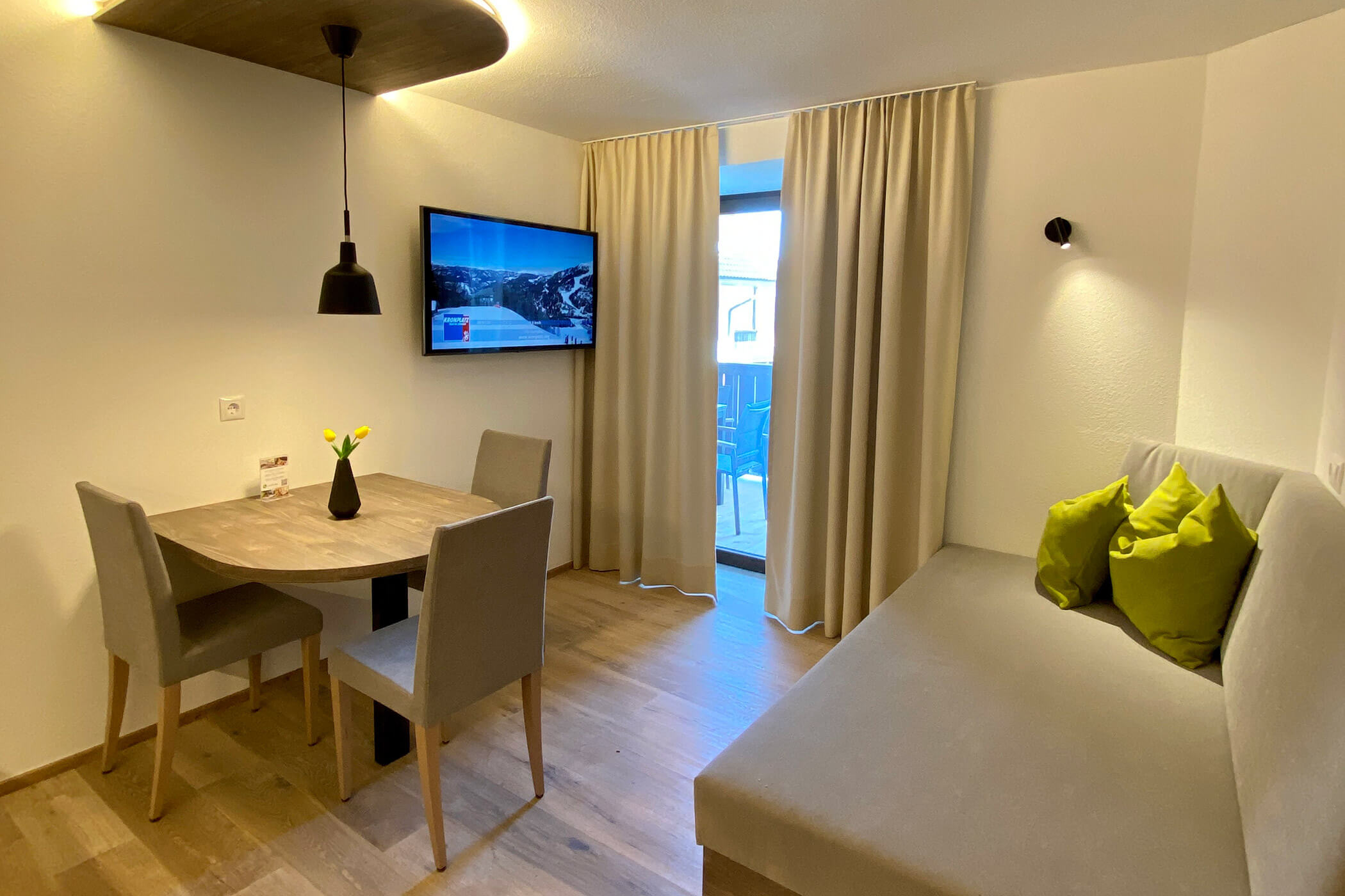 Soggiorno moderno con tavolo, sedie, divano, televisore appeso alla parete e portafinestra con le tende chiuse. - Hotel Oberwiesen