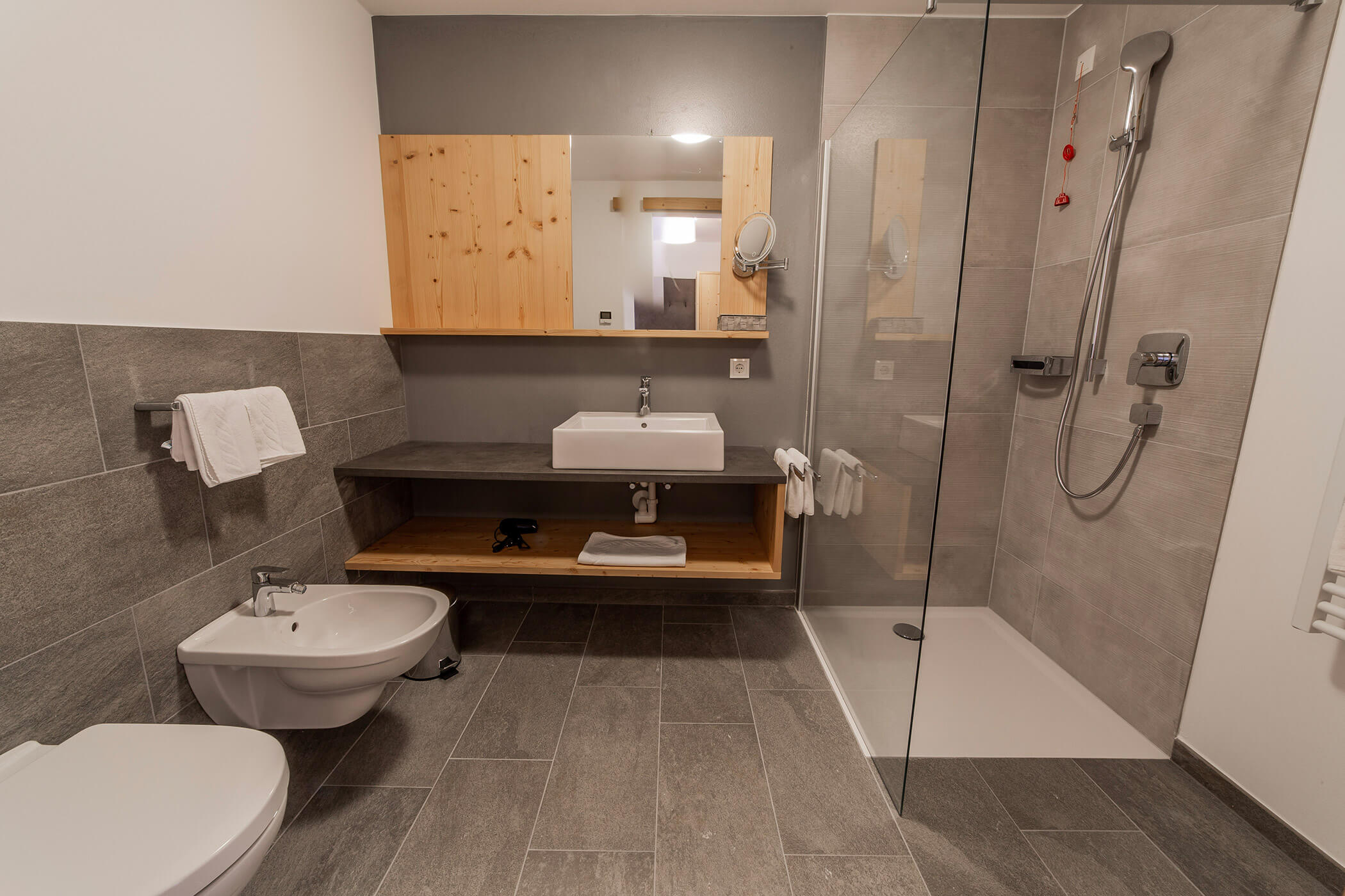 Bagno moderno con piastrelle grigie, lavabo, bidet, WC e box doccia con pareti in vetro. - Hotel Oberwiesen