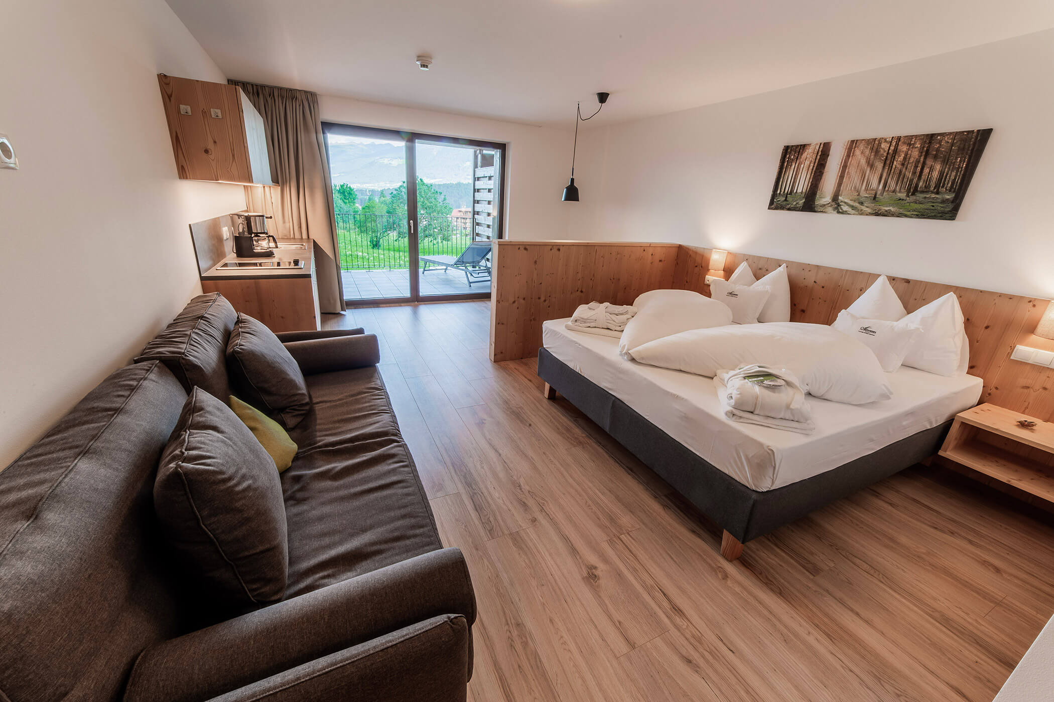 Moderna camera d'albergo con letto, divano, angolo cottura e balcone affacciato sul verde e sugli edifici in lontananza. - Hotel Oberwiesen