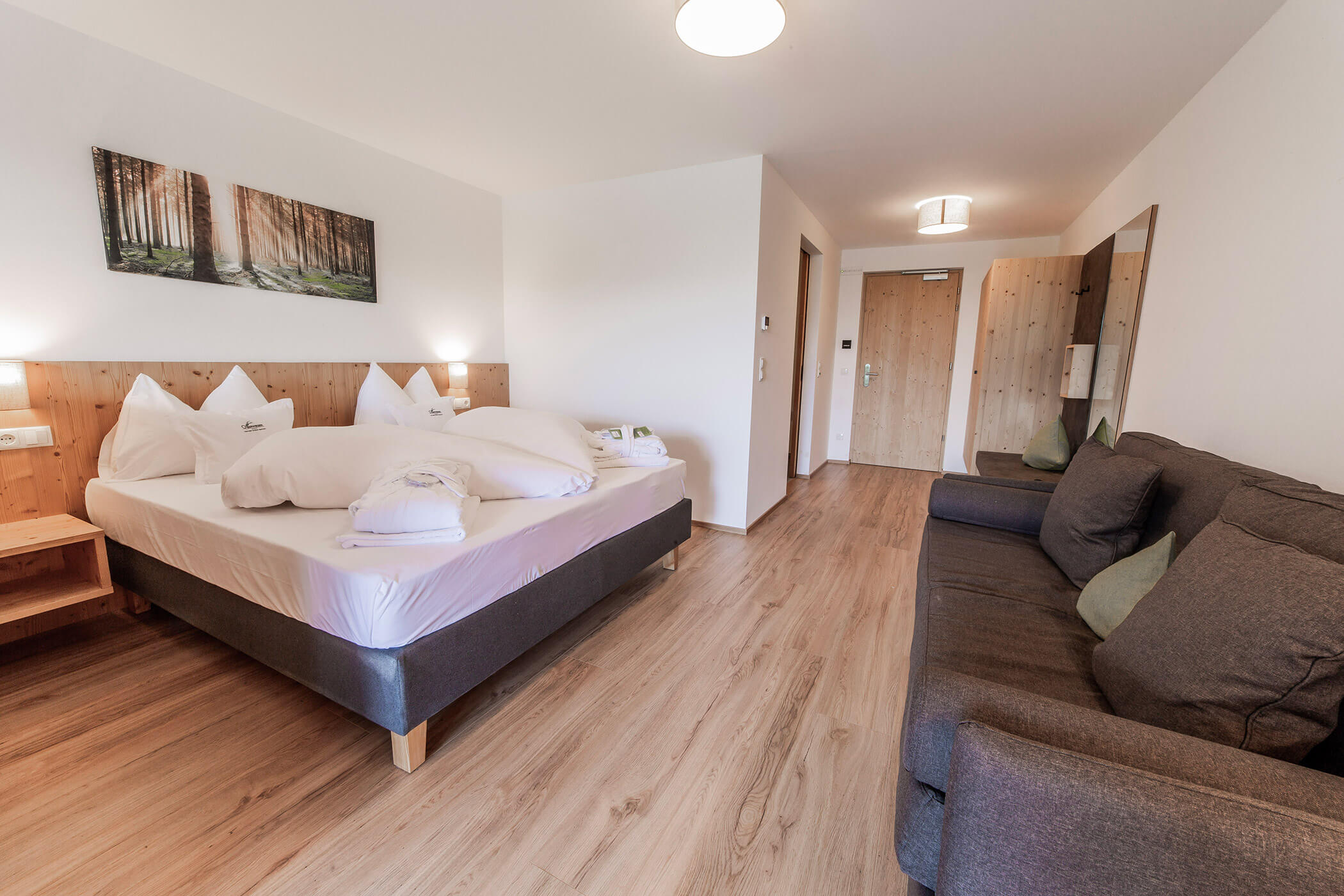 Camera d'albergo moderna con letto matrimoniale, divano, pavimento in legno e un quadro raffigurante un bosco appeso alla parete. - Hotel Oberwiesen