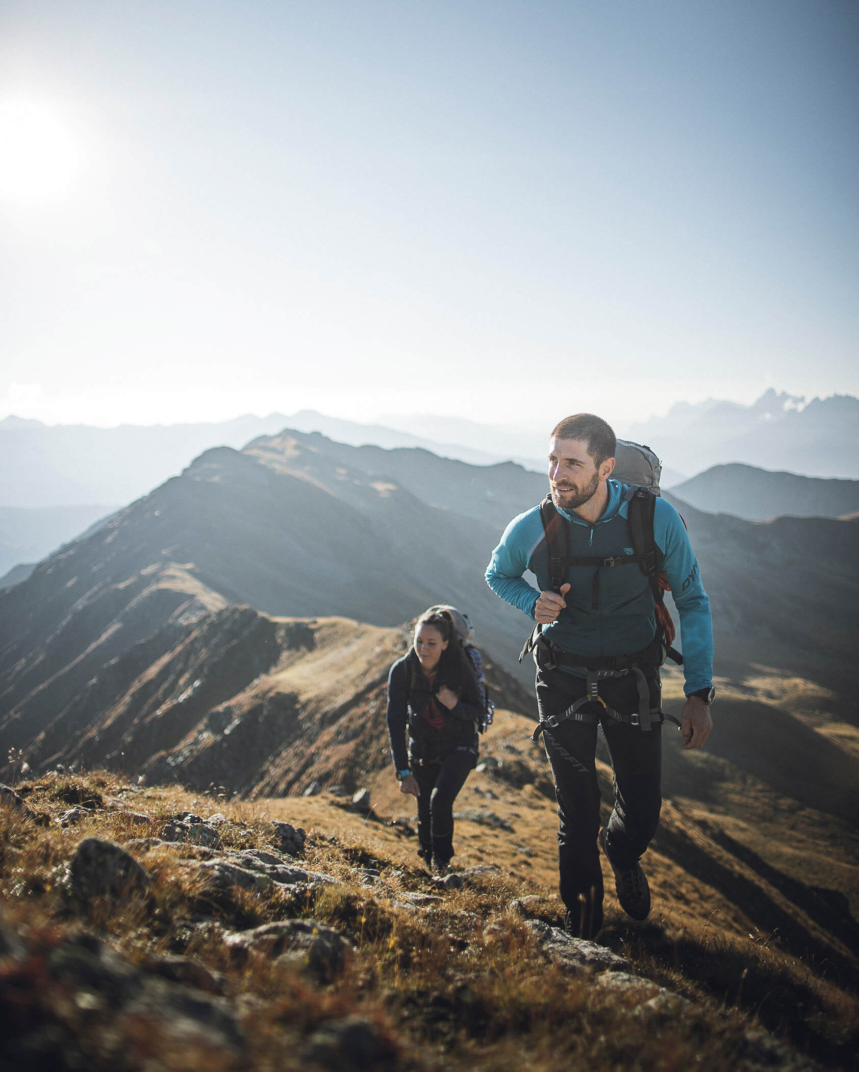 Two people hiking up a grassy mountain slope with distant peaks and sunlight in the background. - Hotel Oberwiesen