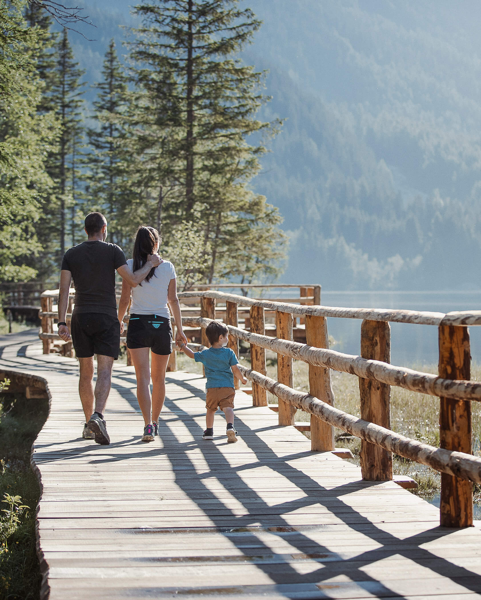 A family of three walks on a wooden path beside a lake, surrounded by trees and mountains. - Hotel Oberwiesen