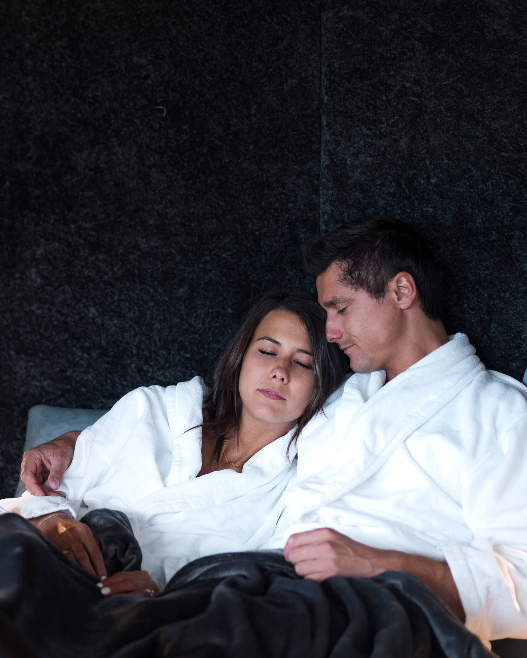 A couple in white robes sits close together, eyes closed, relaxing under a dark blanket. - Hotel Oberwiesen