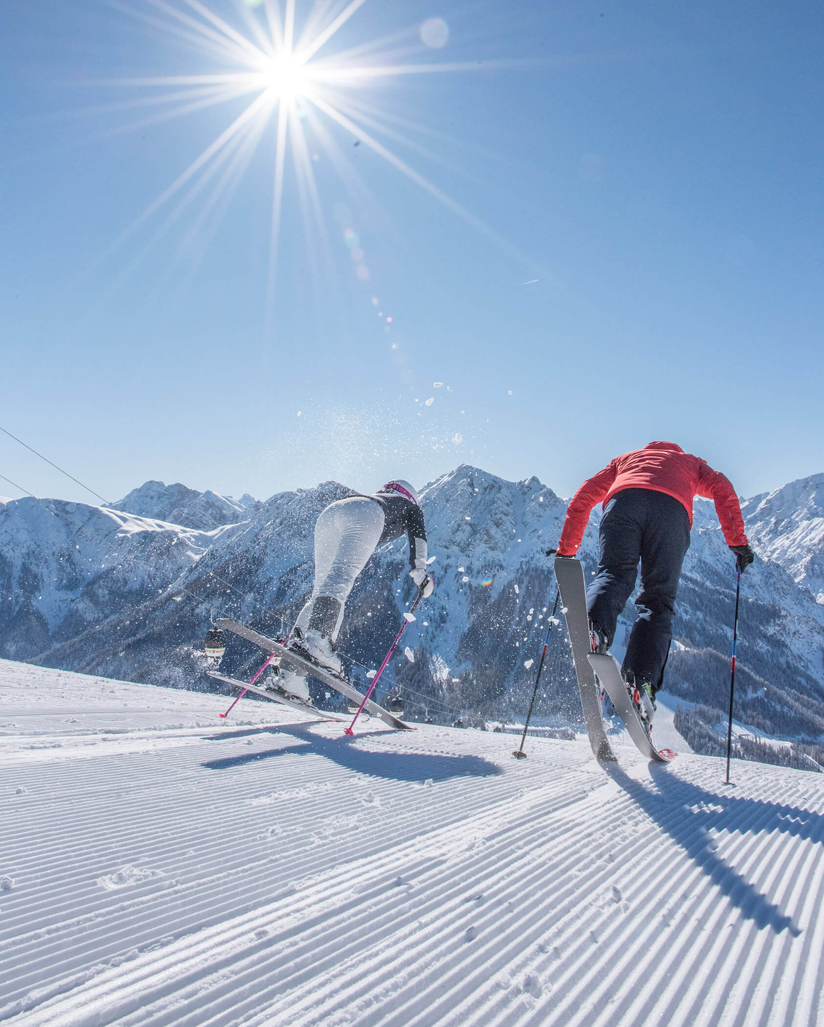 Two skiers push off on a sunny slope, snow-capped mountains and blue sky in the background. - Hotel Oberwiesen