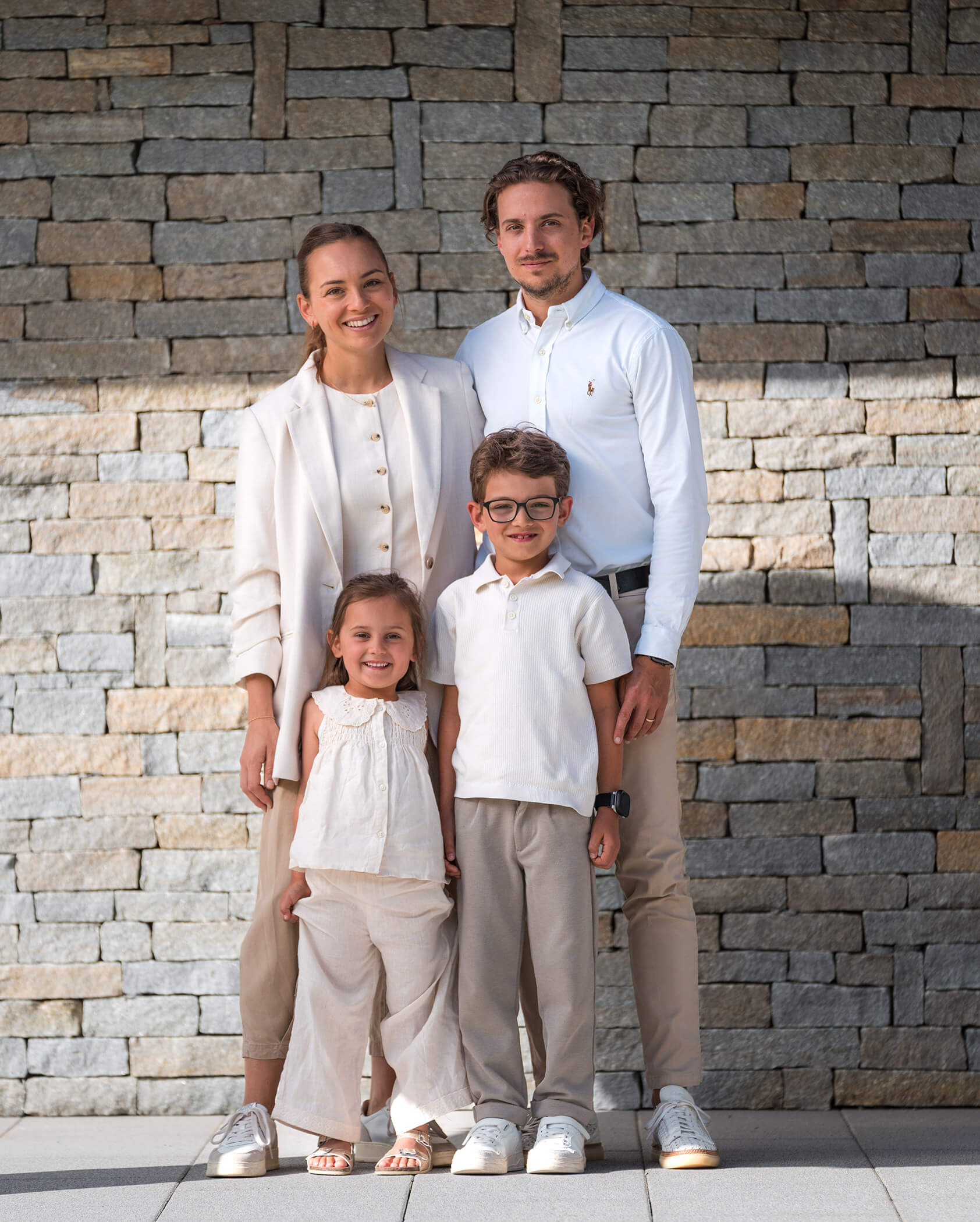 A family of four, all dressed in light clothing, stands smiling in front of a stone wall. - Hotel Oberwiesen