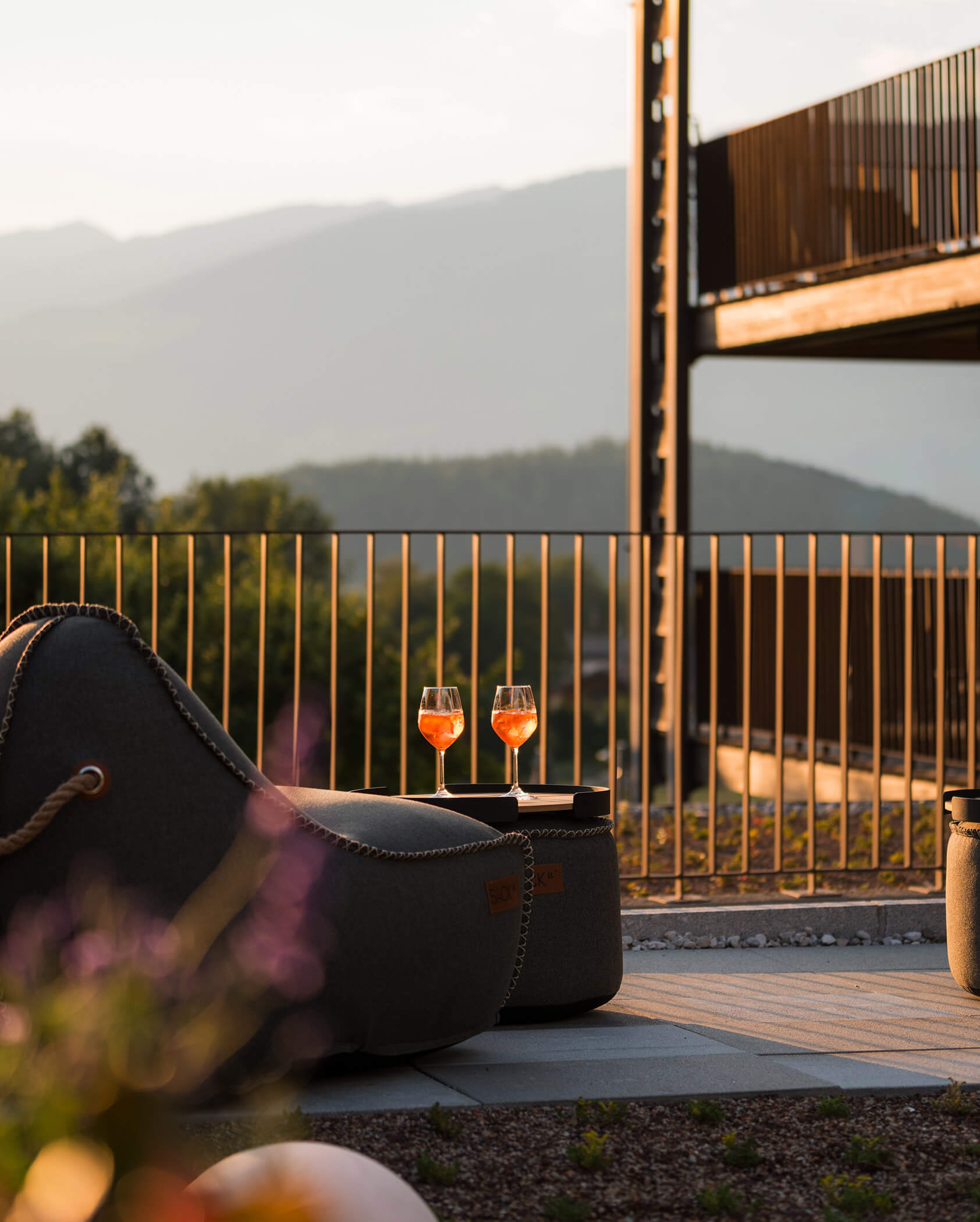 Due bicchieri di vino arancione sono appoggiati su un tavolino accanto a una poltrona, con le montagne sullo sfondo al tramonto. - Hotel Oberwiesen