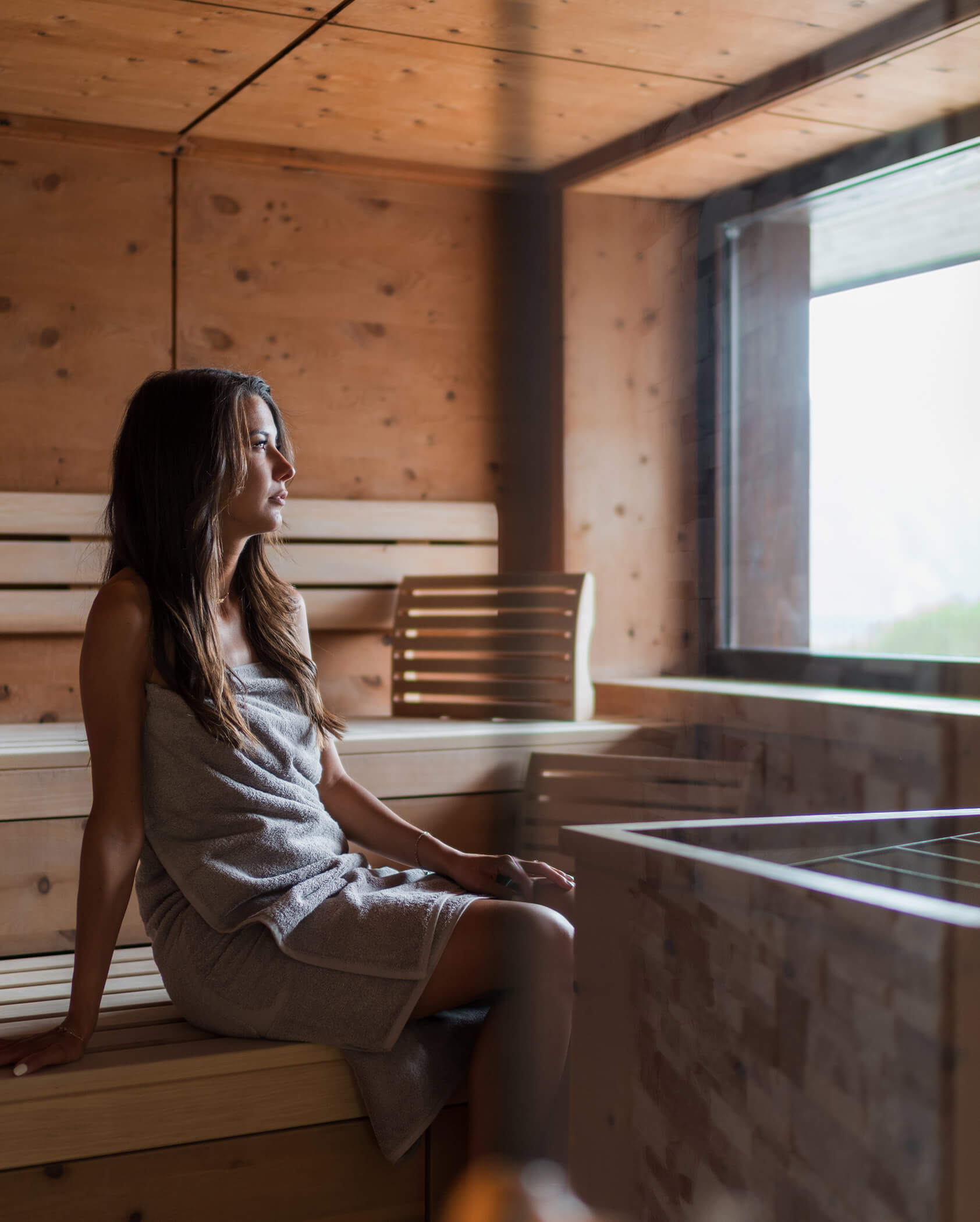Eine Frau in einem Handtuch sitzt auf einer Holzbank in einer Sauna und schaut aus einem Fenster, durch das Tageslicht hereinfällt. - Hotel Oberwiesen