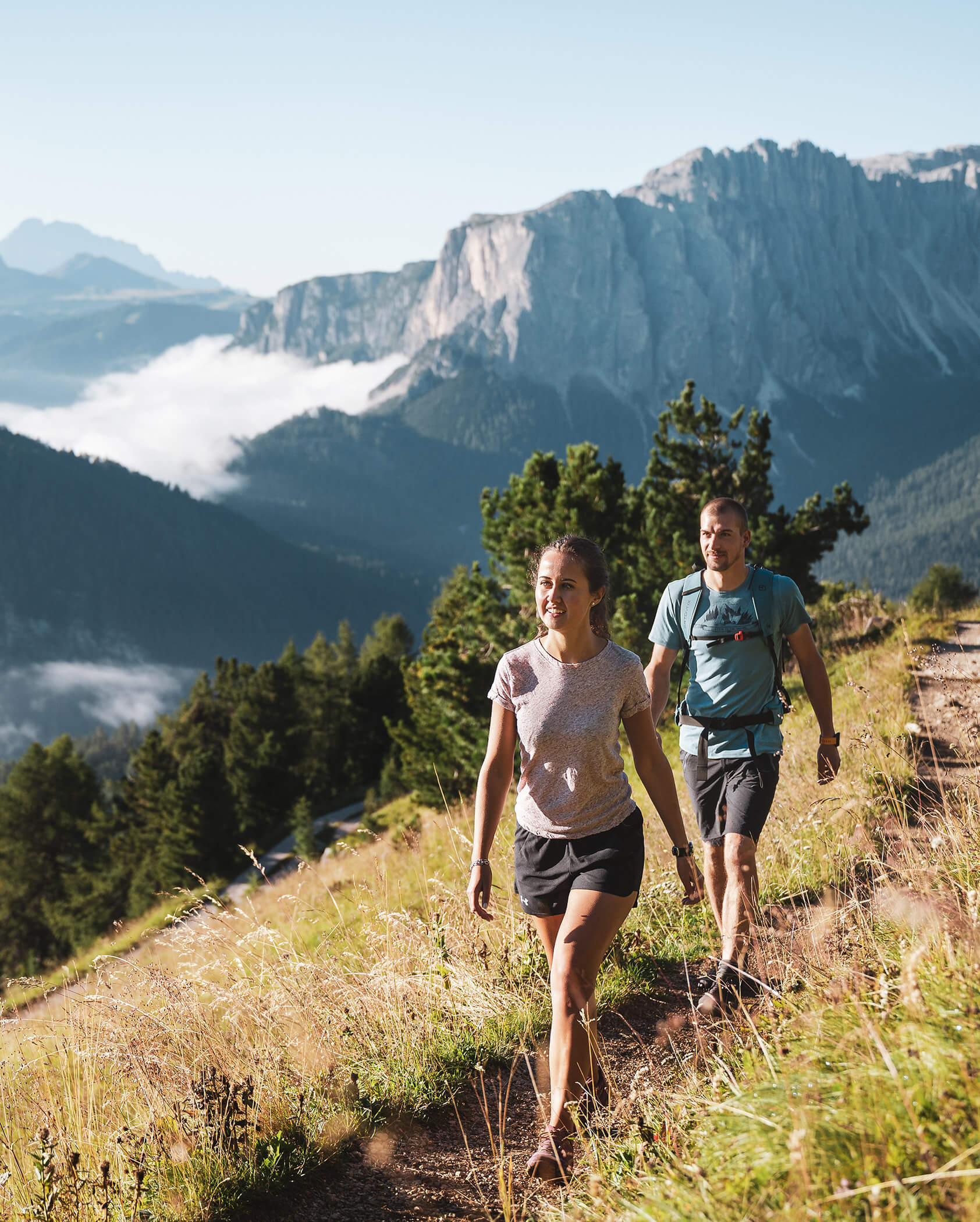 Zwei Menschen wandern auf einem grasbewachsenen Bergpfad, im Hintergrund sind hohe Felsen und Bäume zu sehen. - Hotel Oberwiesen