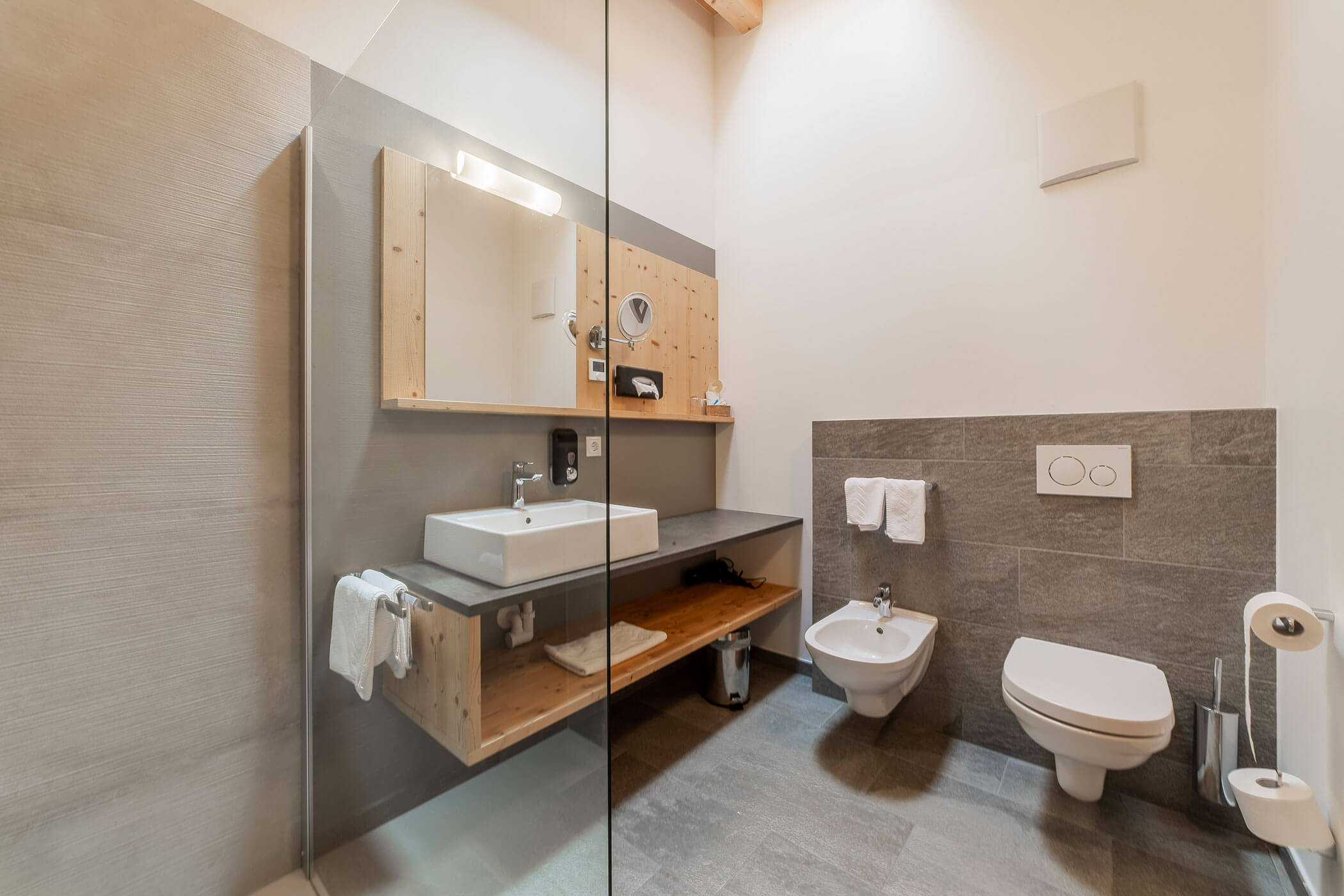 Bagno moderno con lavandino, bidet, WC, asciugamani, dettagli in legno e pavimenti e pareti rivestiti di piastrelle grigie. - Hotel Oberwiesen