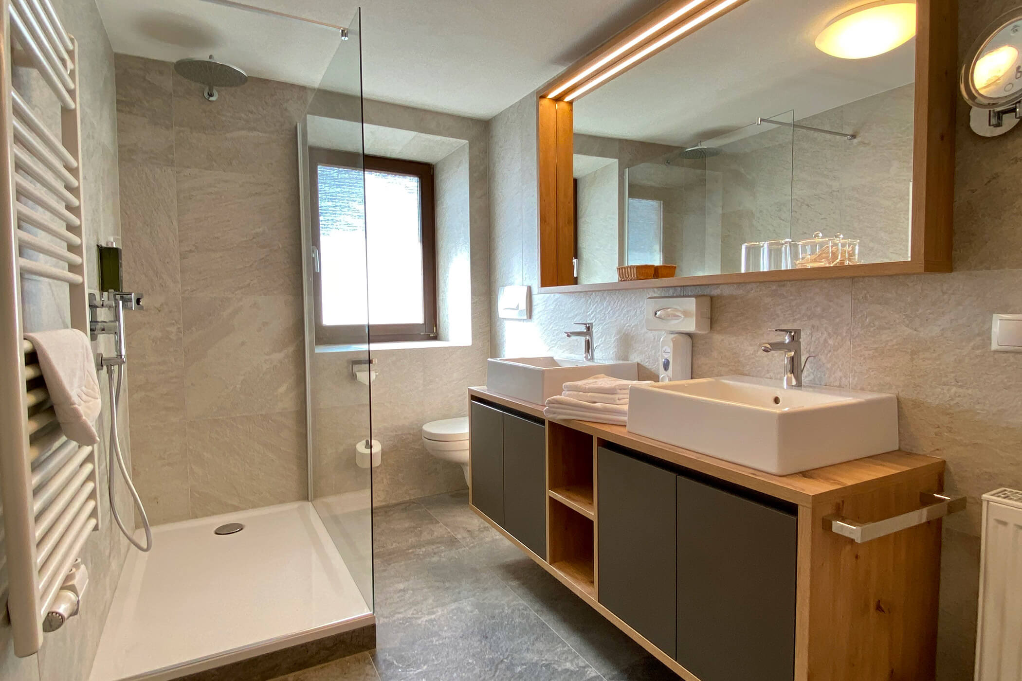 Bagno moderno con box doccia in vetro, doppio lavabo, un grande specchio e luce naturale che entra dalla finestra. - Hotel Oberwiesen