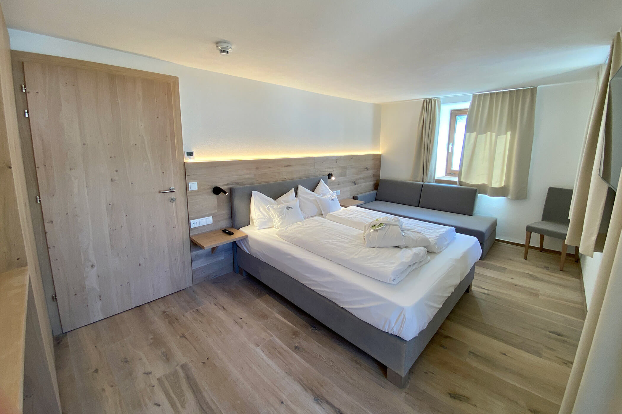 Una camera d'albergo ordinata con un letto matrimoniale, un divano grigio, una poltrona, una porta in legno e un pavimento in legno chiaro. - Hotel Oberwiesen