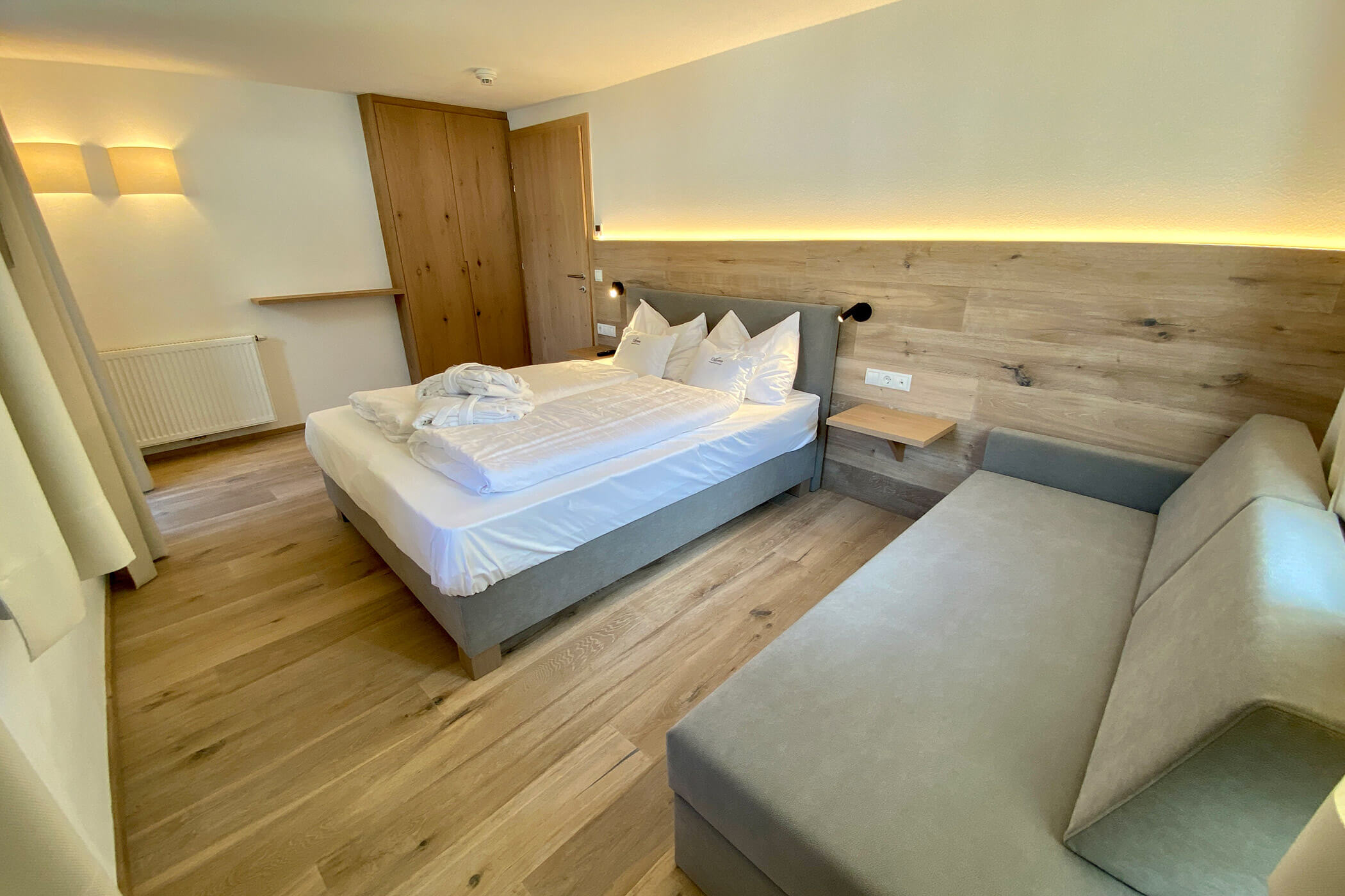 Camera d'albergo moderna con letto matrimoniale, divano, pavimento in legno e illuminazione soffusa sulla parete dietro la testata del letto. - Hotel Oberwiesen
