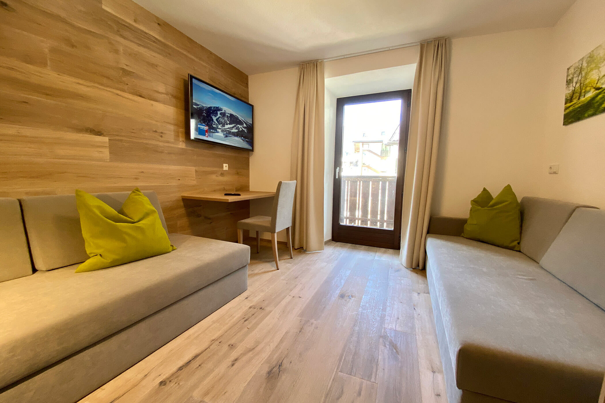 Piccola stanza moderna con due divani, cuscini verdi, una TV a parete, una scrivania, una sedia e una porta che dà sul balcone. - Hotel Oberwiesen