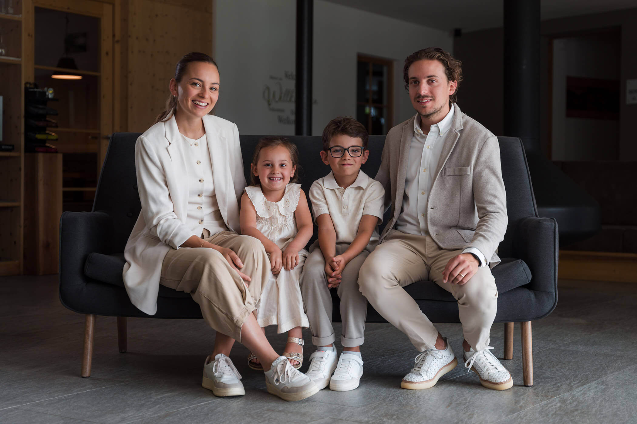 Eine vierköpfige Familie sitzt auf einem dunklen Sofa, alle lächeln und tragen aufeinander abgestimmte helle Kleidung. - Hotel Oberwiesen
