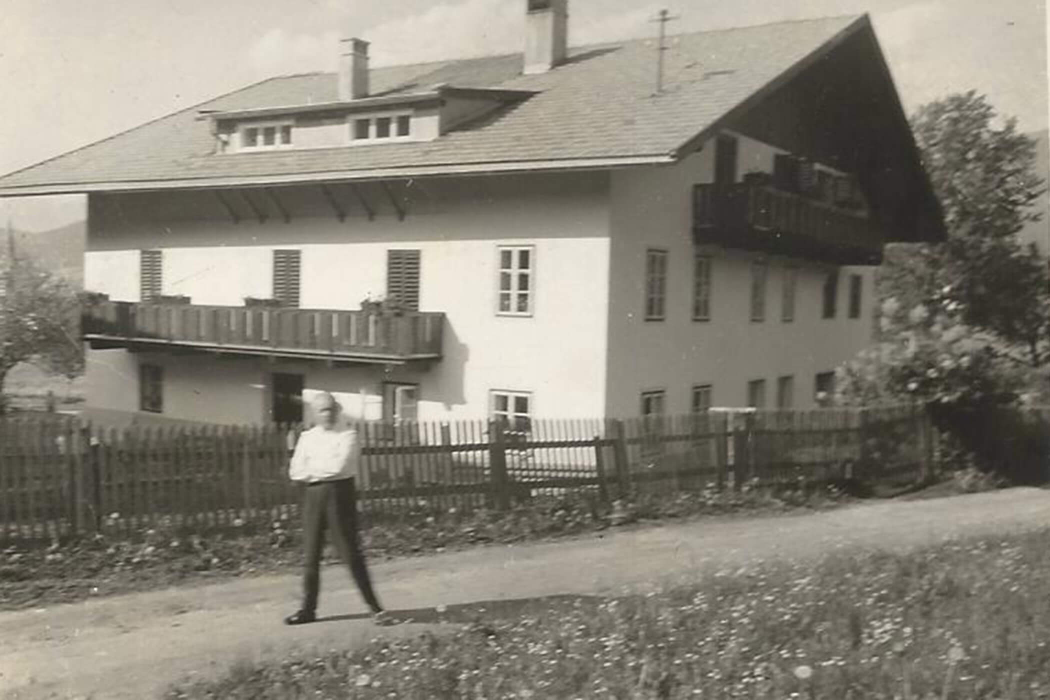 Eine Person steht auf einem Weg vor einem großen Haus mit Balkonen, einem Holzzaun und Bäumen. - Hotel Oberwiesen