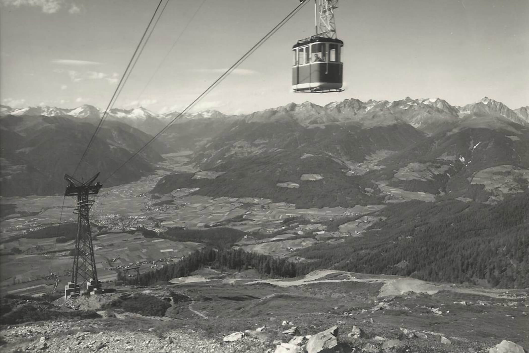 Eine Seilbahn fährt unter klarem Himmel über eine bergige Landschaft mit Tälern und fernen Gipfeln hinauf. - Hotel Oberwiesen