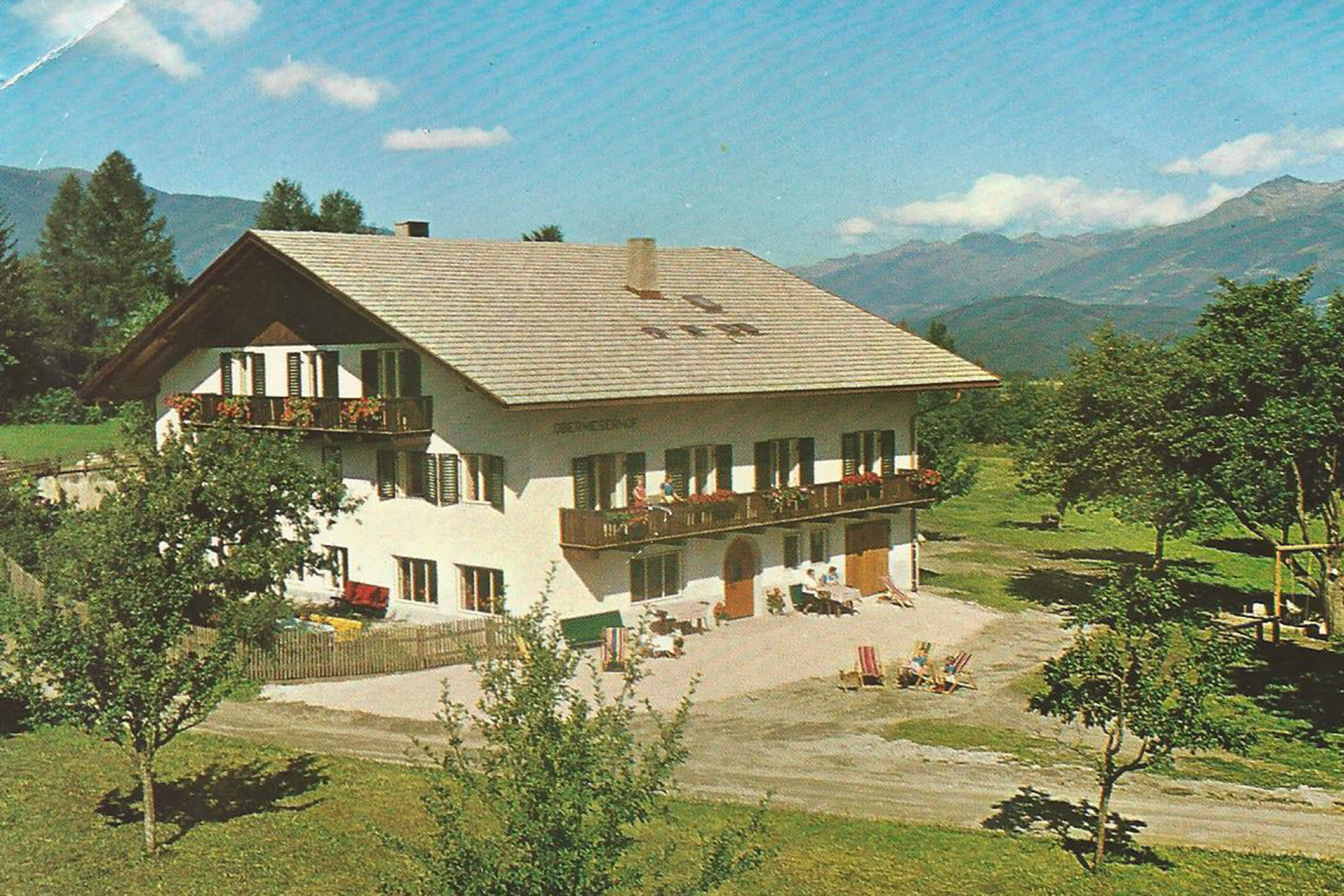 Ein großes weißes Haus mit Balkonen, umgeben von Bäumen und Bergen unter einem strahlend blauen Himmel. - Hotel Oberwiesen