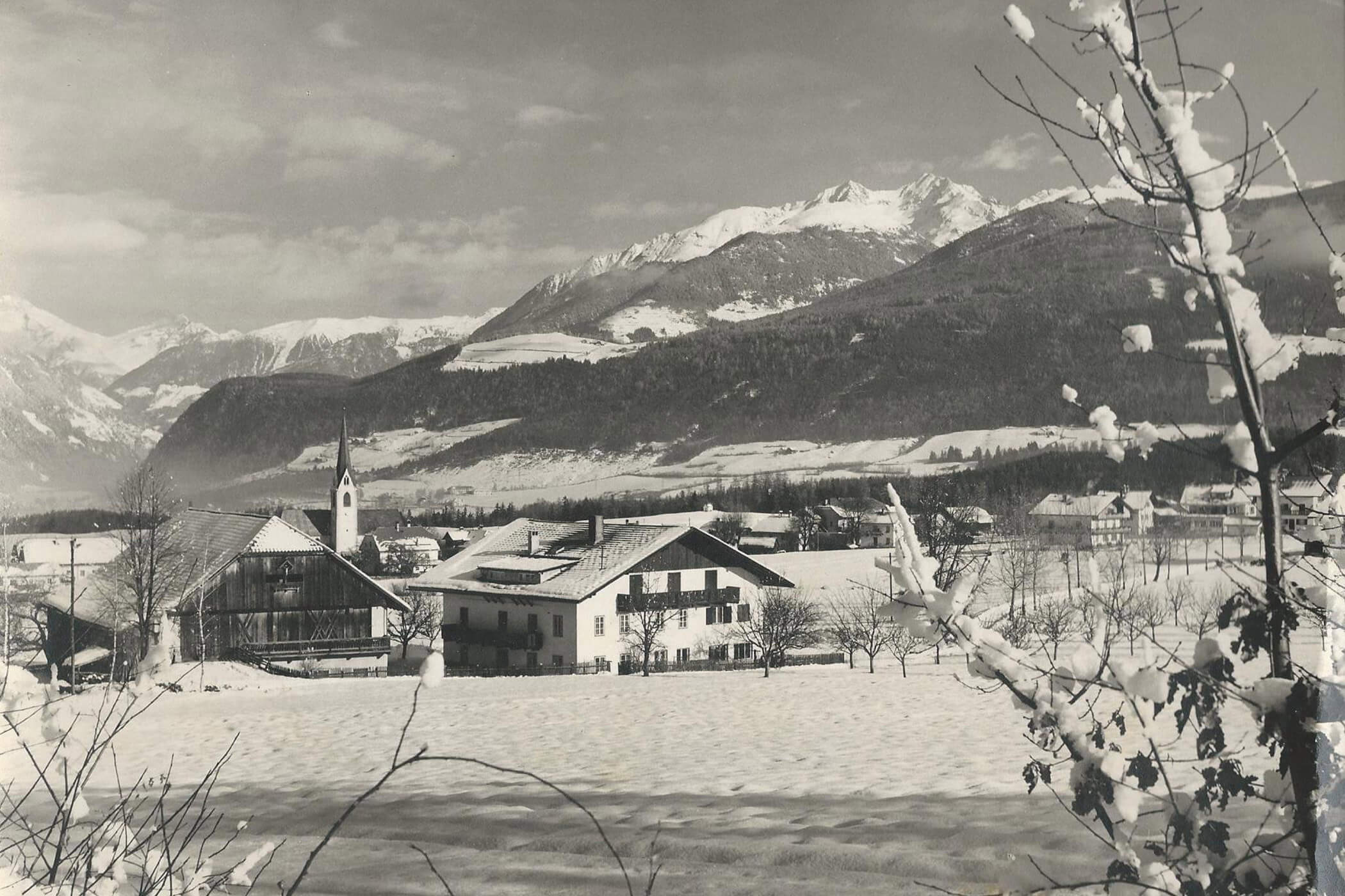 Ein verschneites Dorf mit Kirchturm, Häusern und Bergen im Hintergrund unter einem bewölkten Himmel. - Hotel Oberwiesen