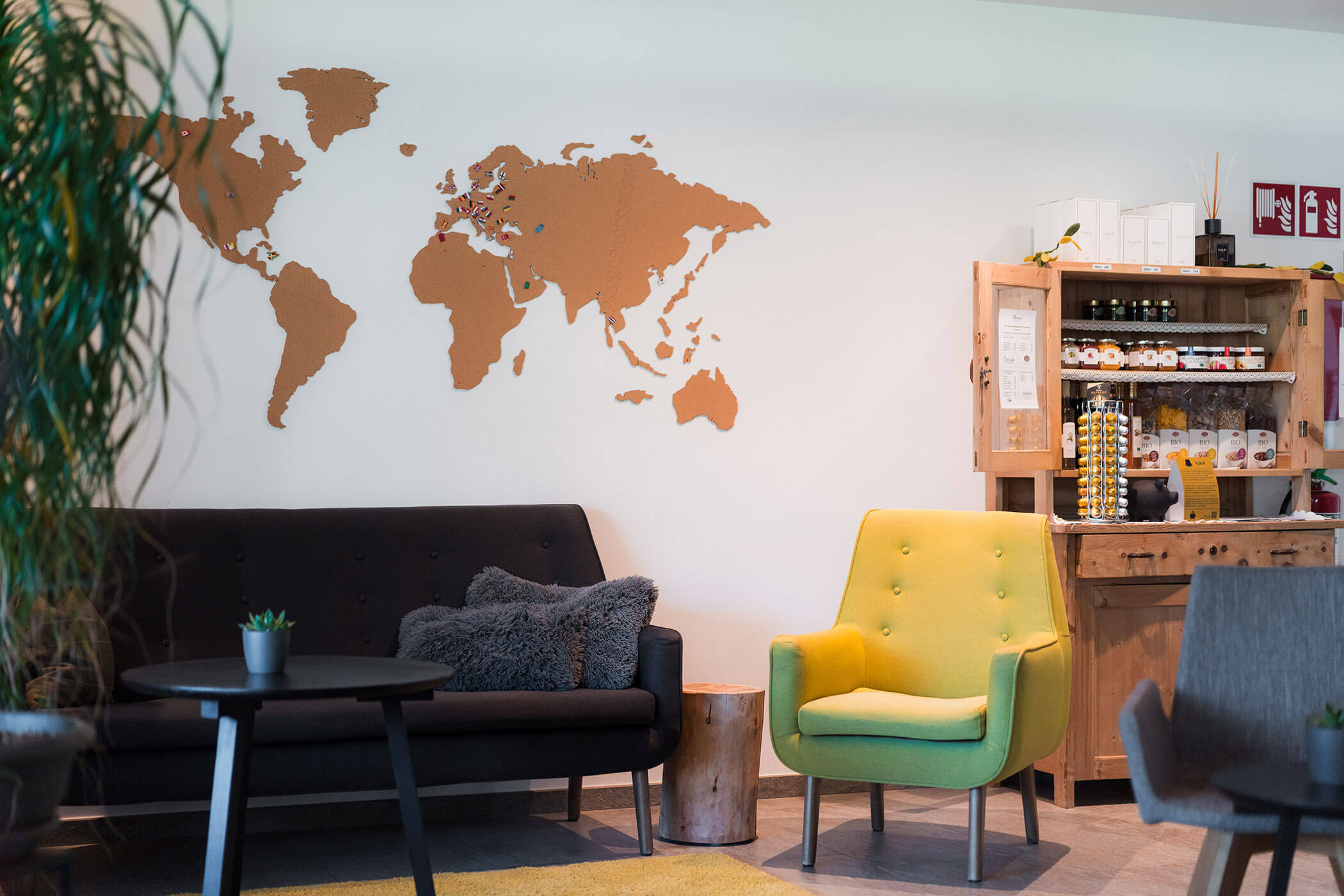 Moderne Lounge mit einem schwarzen Sofa, einem gelben Sessel, einer Wanddekoration in Form einer Weltkarte und einem Holzschrank mit Vorräten. - Hotel Oberwiesen