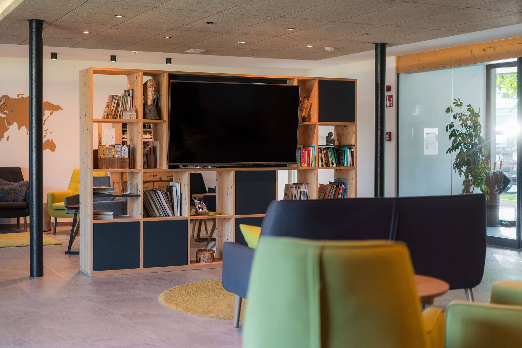 Moderne Lounge mit Bücherregalen, Fernseher, bunten Stühlen und großen Fenstern, durch die Tageslicht hereinströmt. - Hotel Oberwiesen