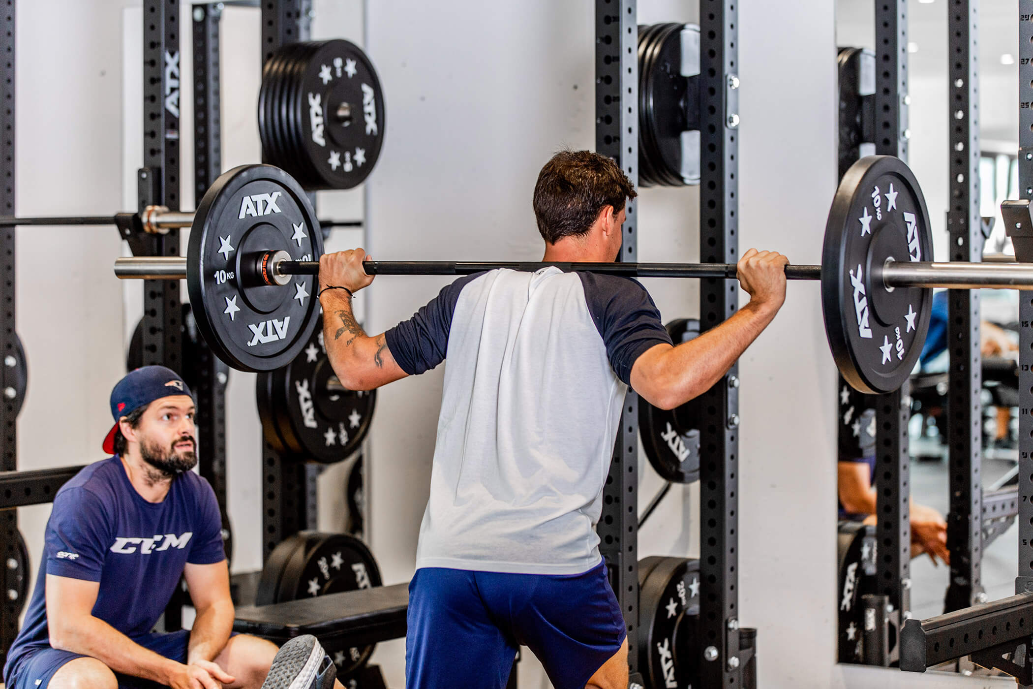 Ein Mann hebt in einem Fitnessstudio eine Langhantel für Kniebeugen, während ein anderer Mann mit einer Mütze in der Nähe sitzt und ihm dabei zusieht. - Hotel Oberwiesen