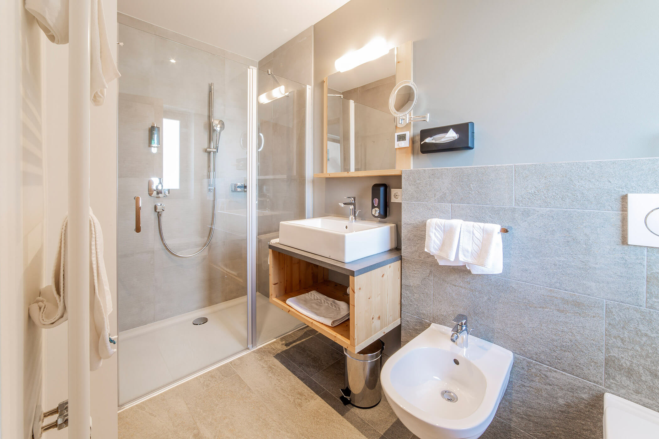 Bagno moderno con cabina doccia, lavabo, bidet, portasciugamani, specchio e pareti rivestite con piastrelle grigio chiaro. - Hotel Oberwiesen