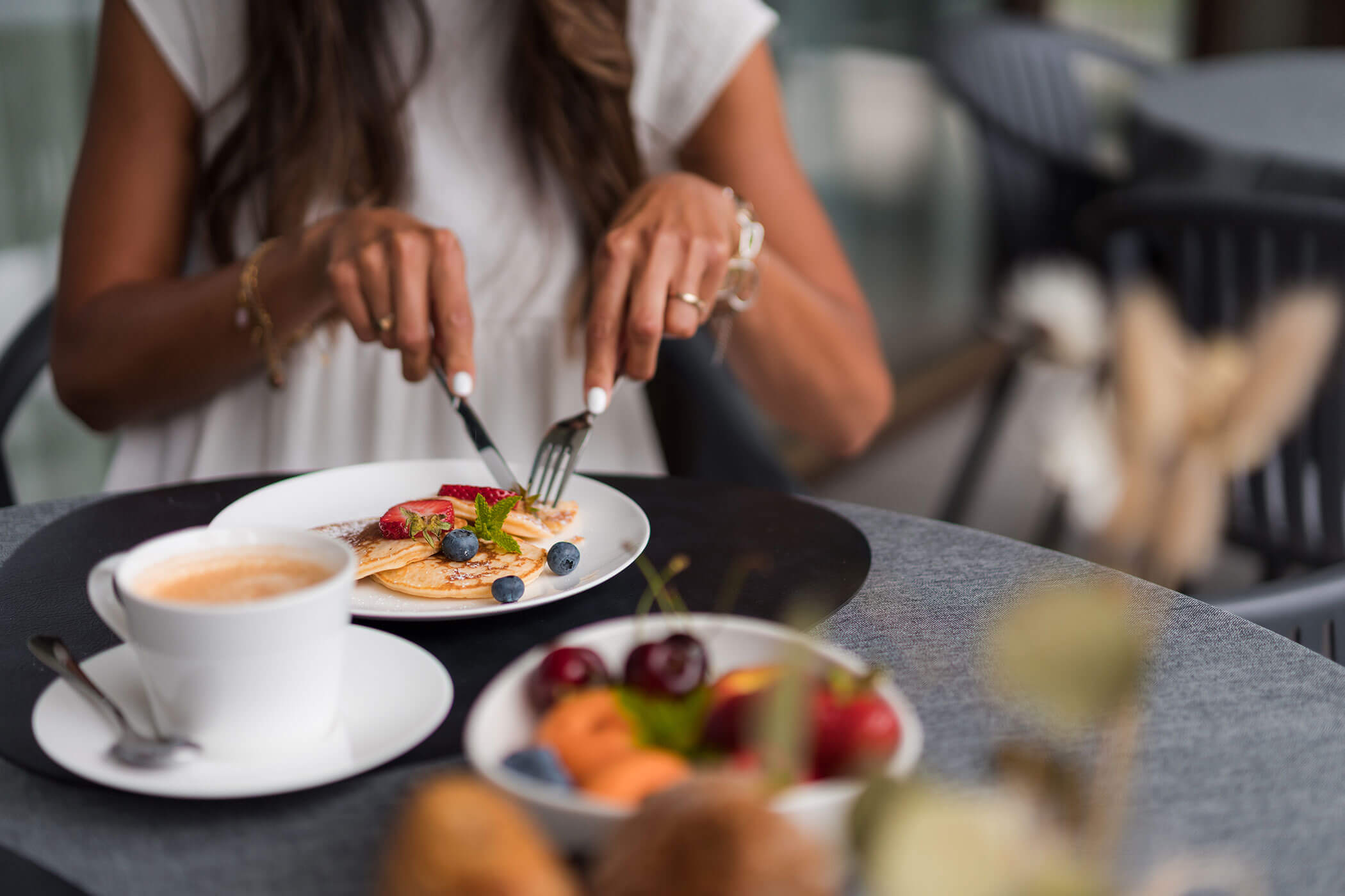 Una donna che taglia dei pancake con frutta su un piatto, con una tazza di caffè e una ciotola di frutta sul tavolo. - Hotel Oberwiesen