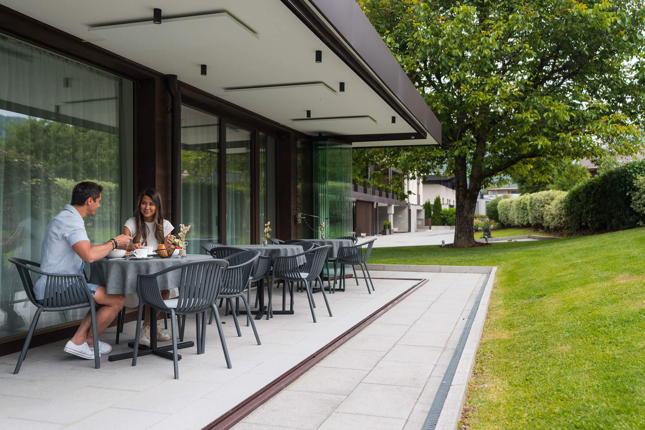 Una coppia è seduta a un tavolo all’aperto, intenta a mangiare e a chiacchierare vicino a un edificio moderno e a un prato verde. - Hotel Oberwiesen