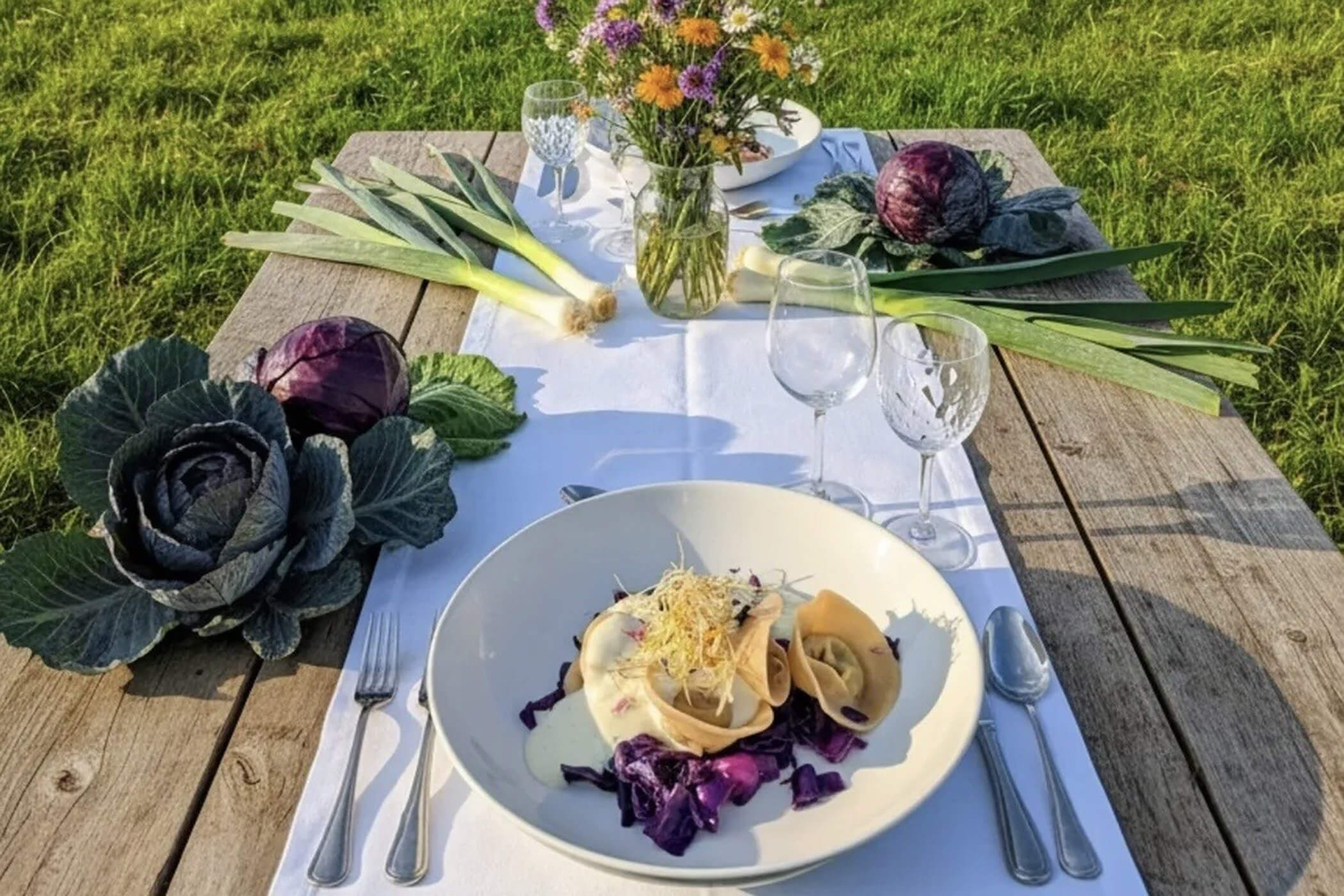 Tavola all’aperto apparecchiata con un piatto di pasta, bicchieri da vino, cavoli, porri e un vaso di fiori di campo. - Hotel Oberwiesen