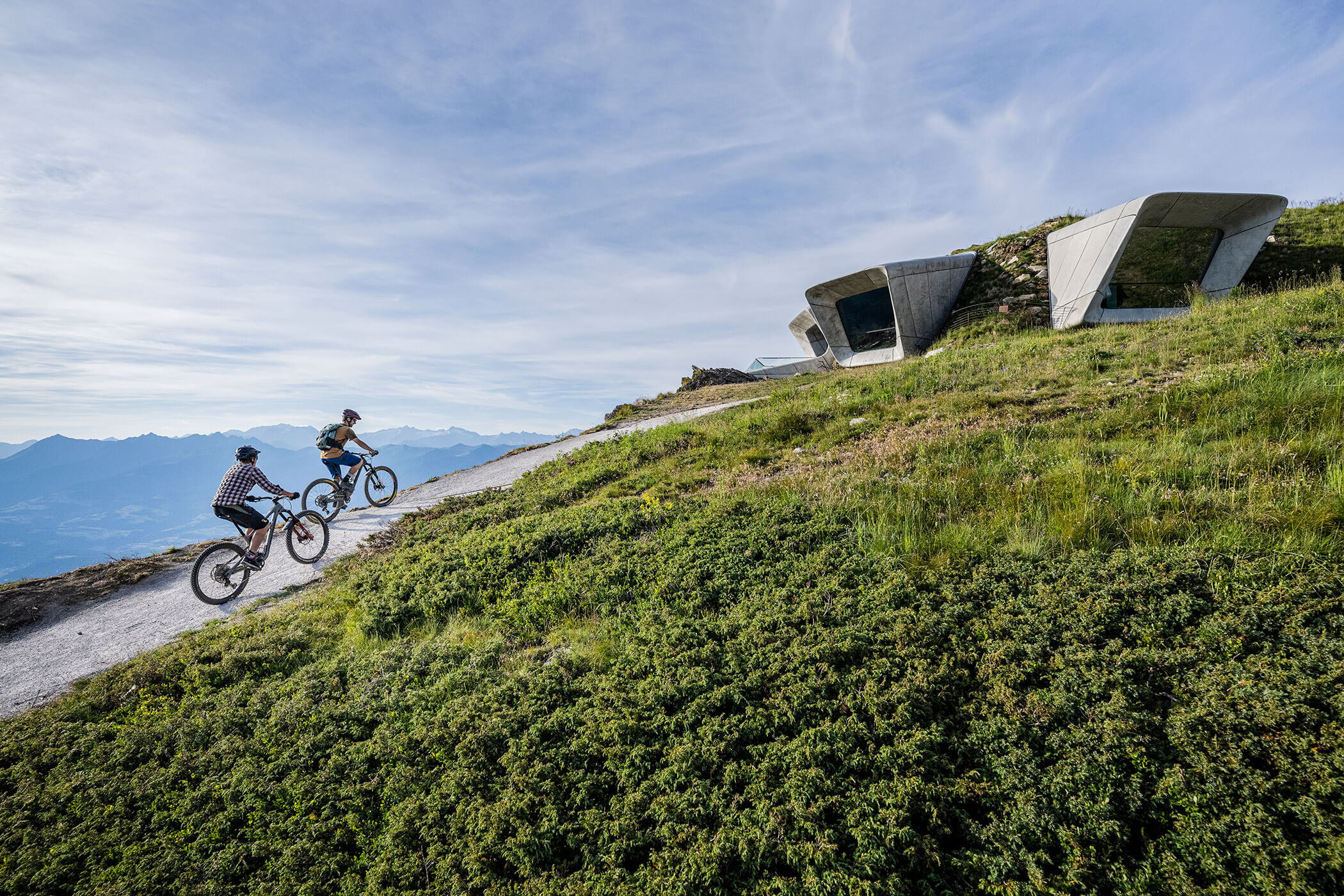 Two people ride bikes uphill on a trail near modern buildings, surrounded by green grass and mountains. - Hotel Oberwiesen