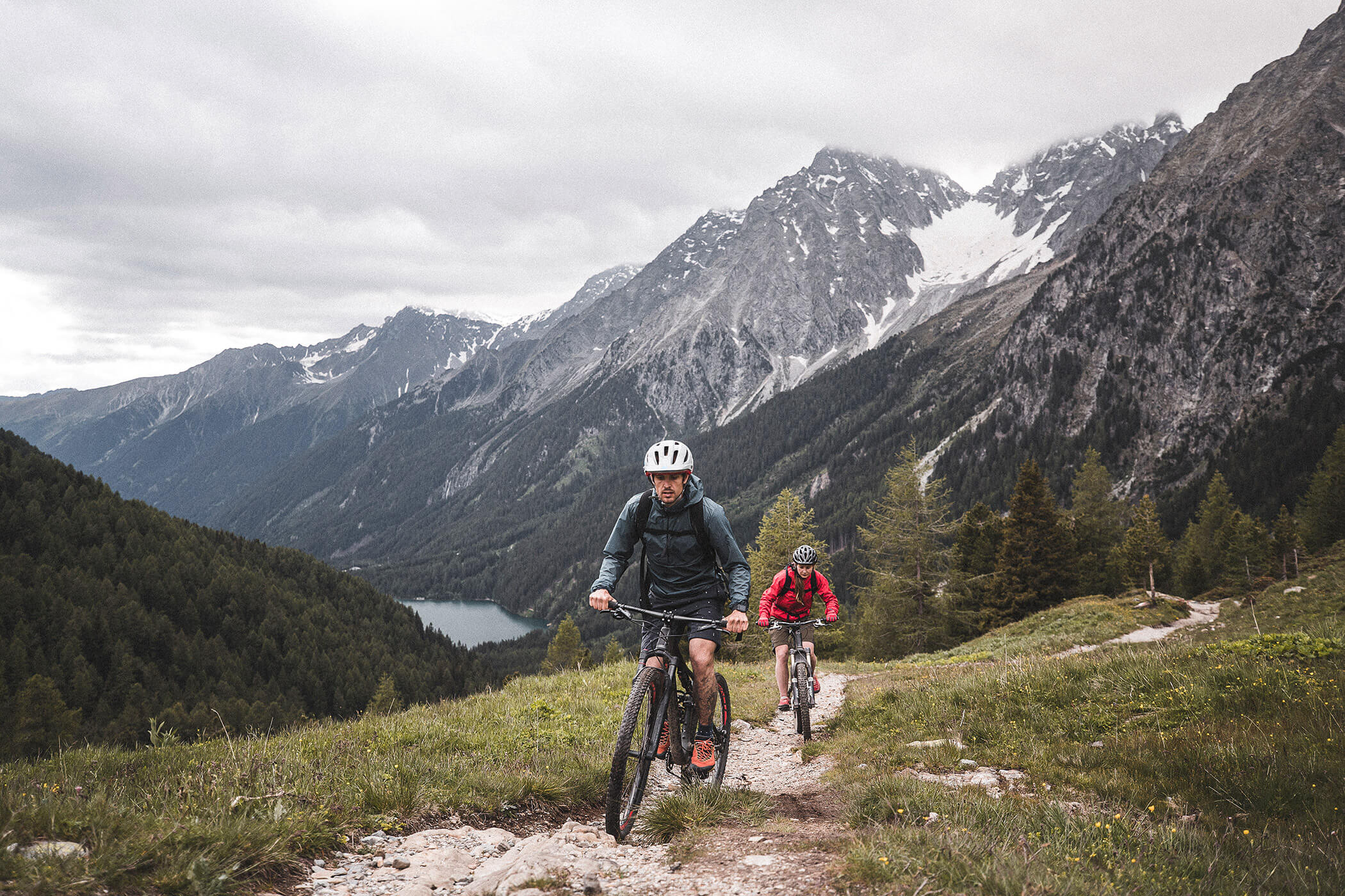 Two people ride mountain bikes on a trail with forested hills and snow-capped mountains in the background. - Hotel Oberwiesen