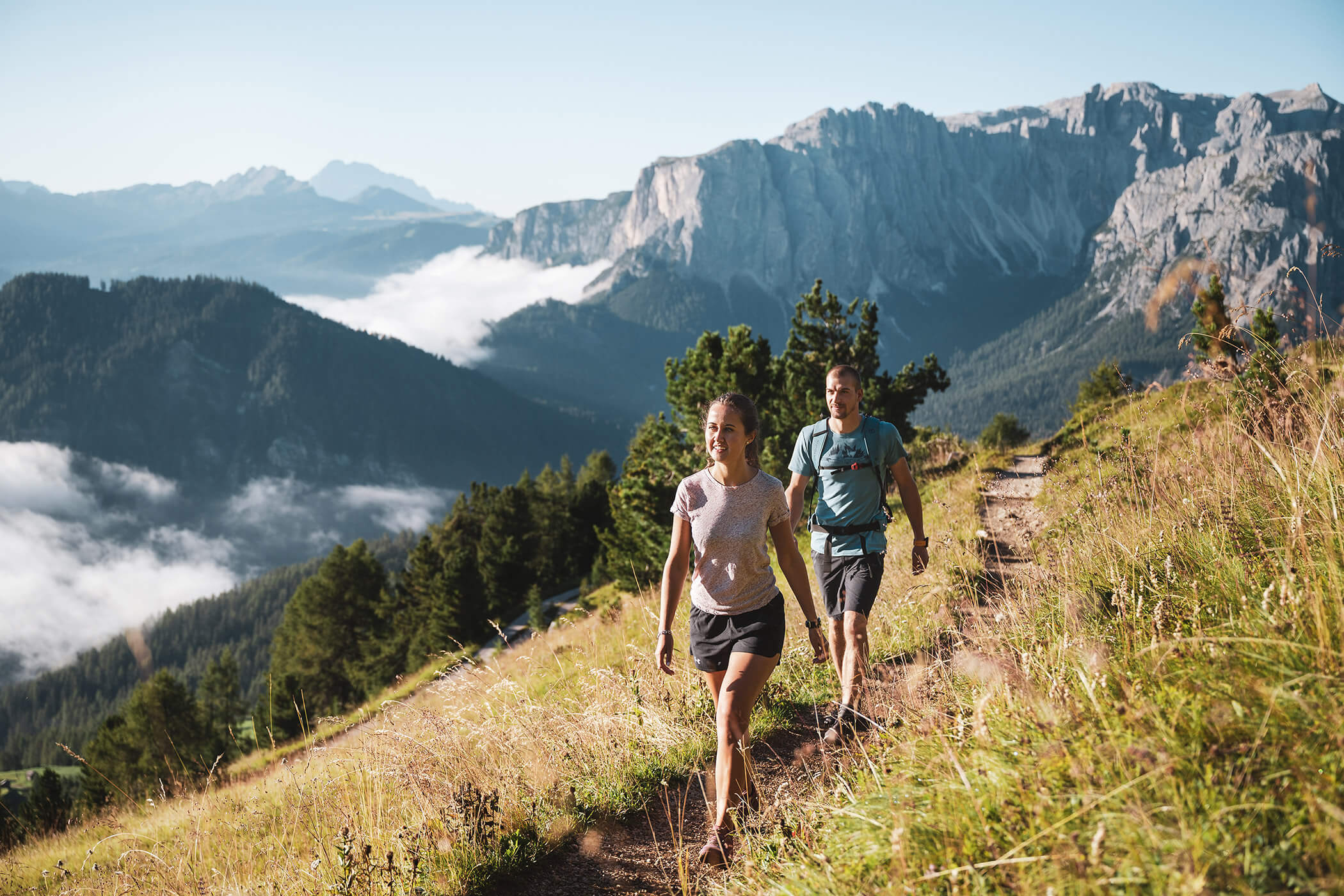 Two people hiking a grassy mountain trail with scenic cliffs and forests in the background under a clear sky. - Hotel Oberwiesen