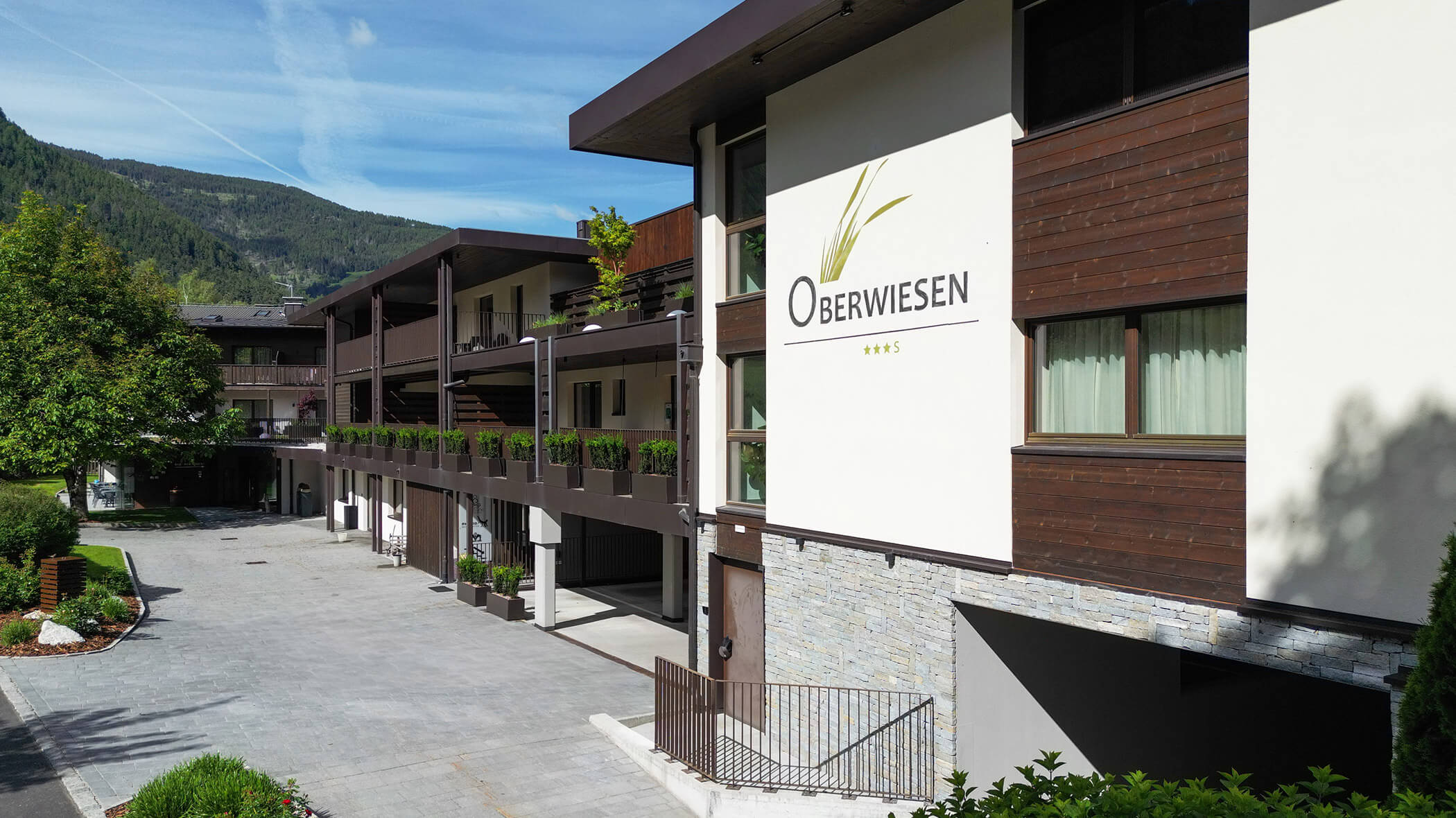 Modern hotel with wooden balconies, greenery, and a sign reading Oberwiesen on the facade. Mountain backdrop. - Hotel Oberwiesen
