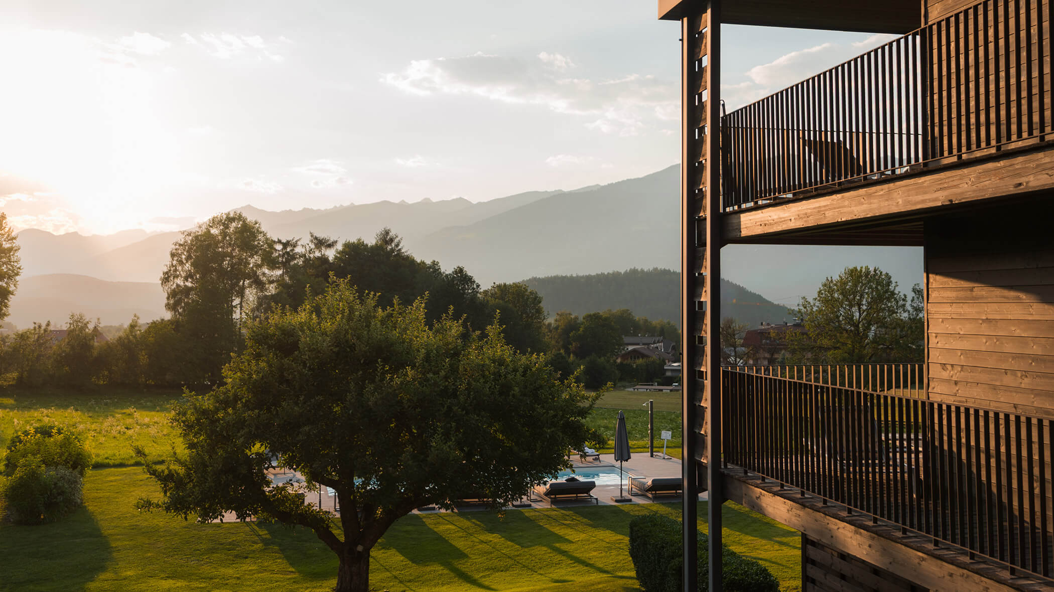 Balconies overlook a green yard with trees, pool, and distant mountains under a hazy sunset sky. - Hotel Oberwiesen