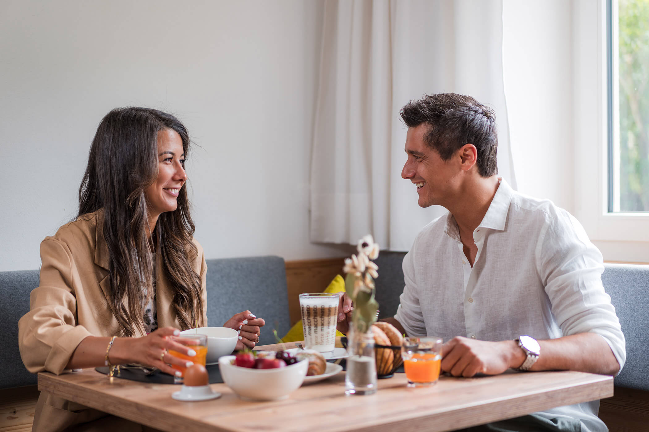 Two people sit at a table with breakfast foods, smiling and talking by a bright window. - Hotel Oberwiesen