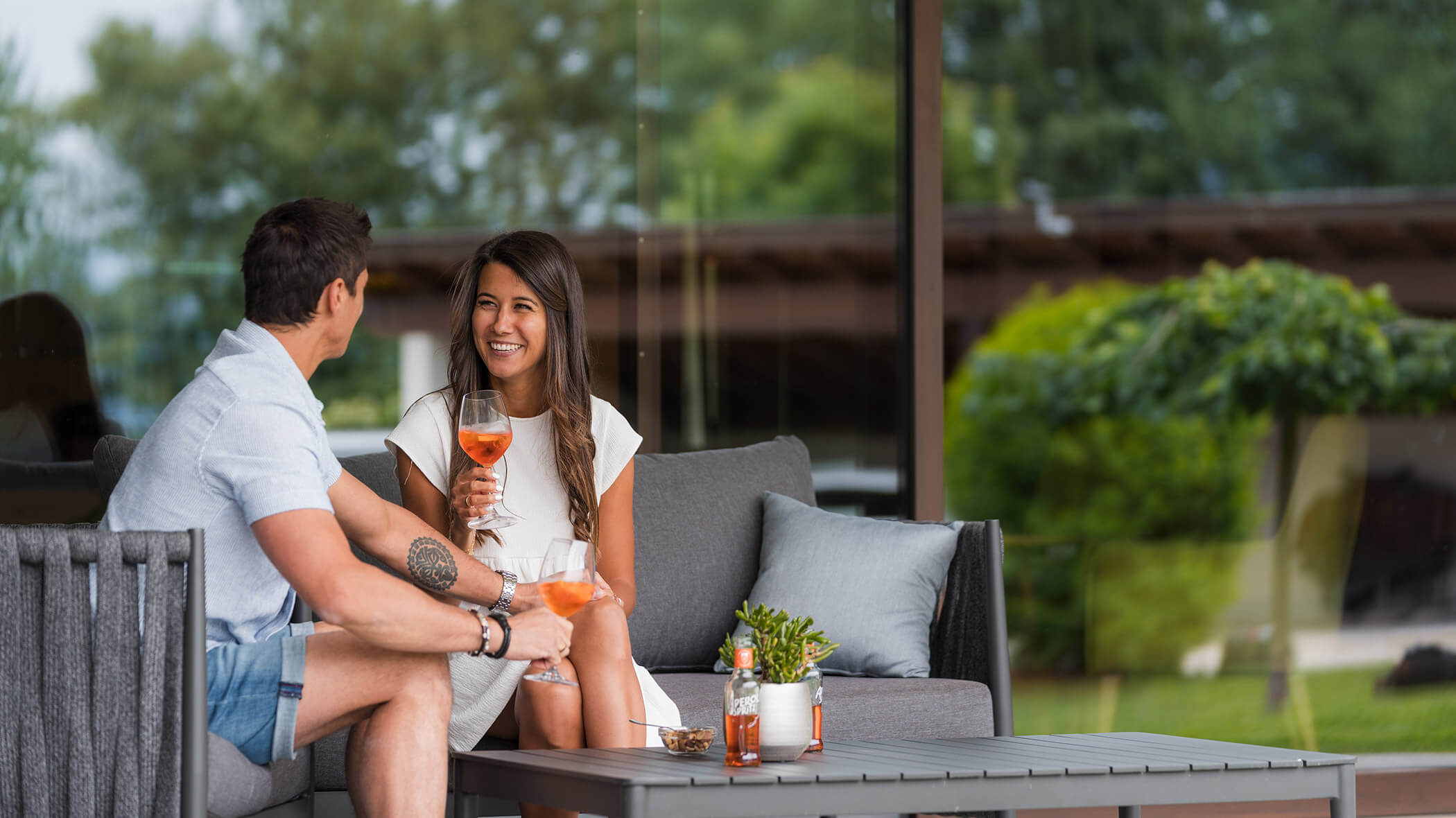 A man and woman sit outside on a sofa, smiling and holding drinks, with greenery in the background. - Hotel Oberwiesen
